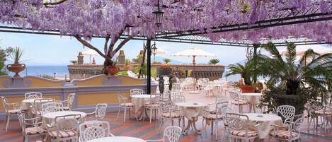 An outdoor patio with white tables and chairs, a view of the sea, and purple flowering trees.