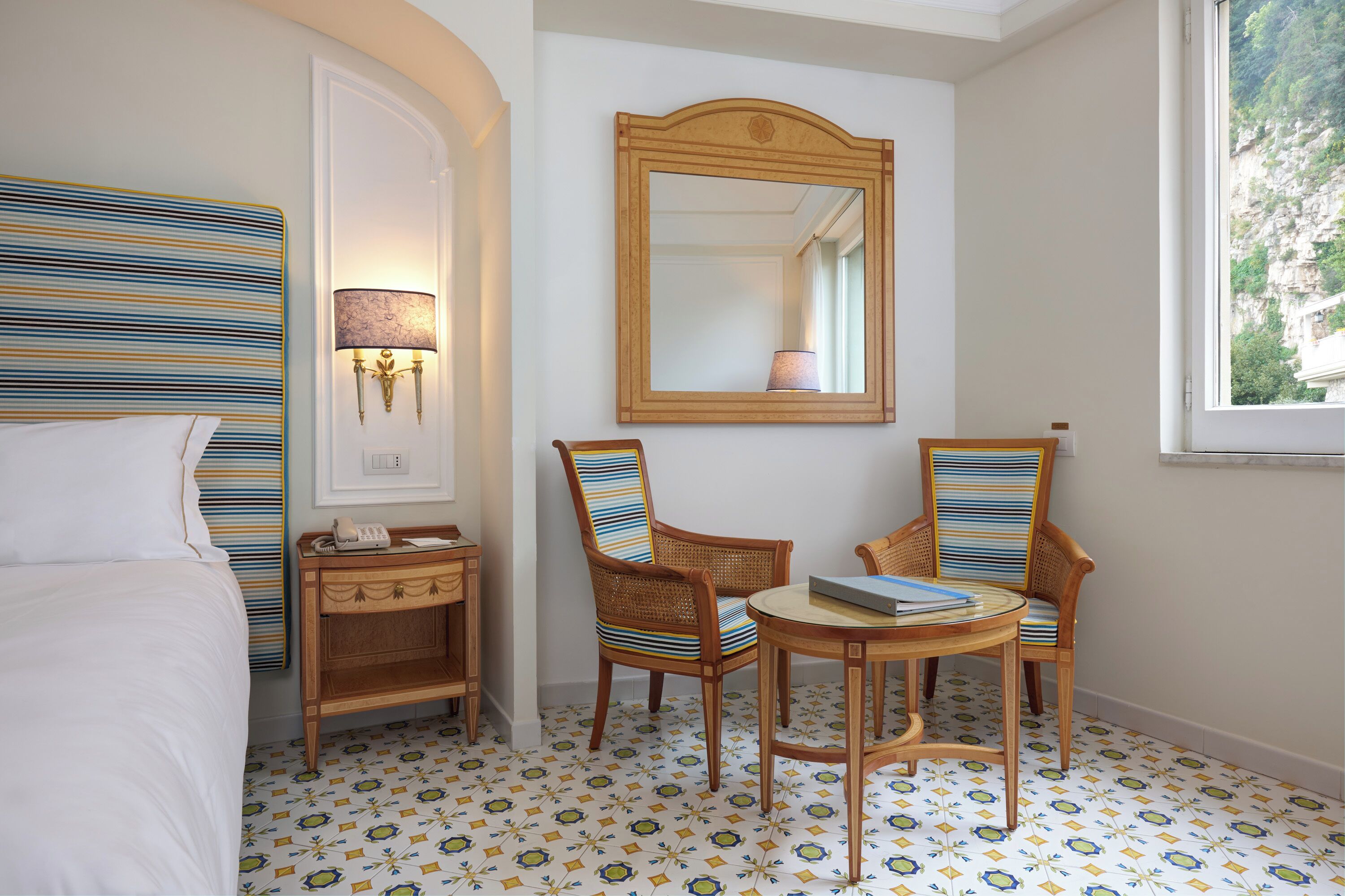 A hotel room with a bed, a desk with a telephone, two chairs, a round table, a mirror, and patterned floor tiles.