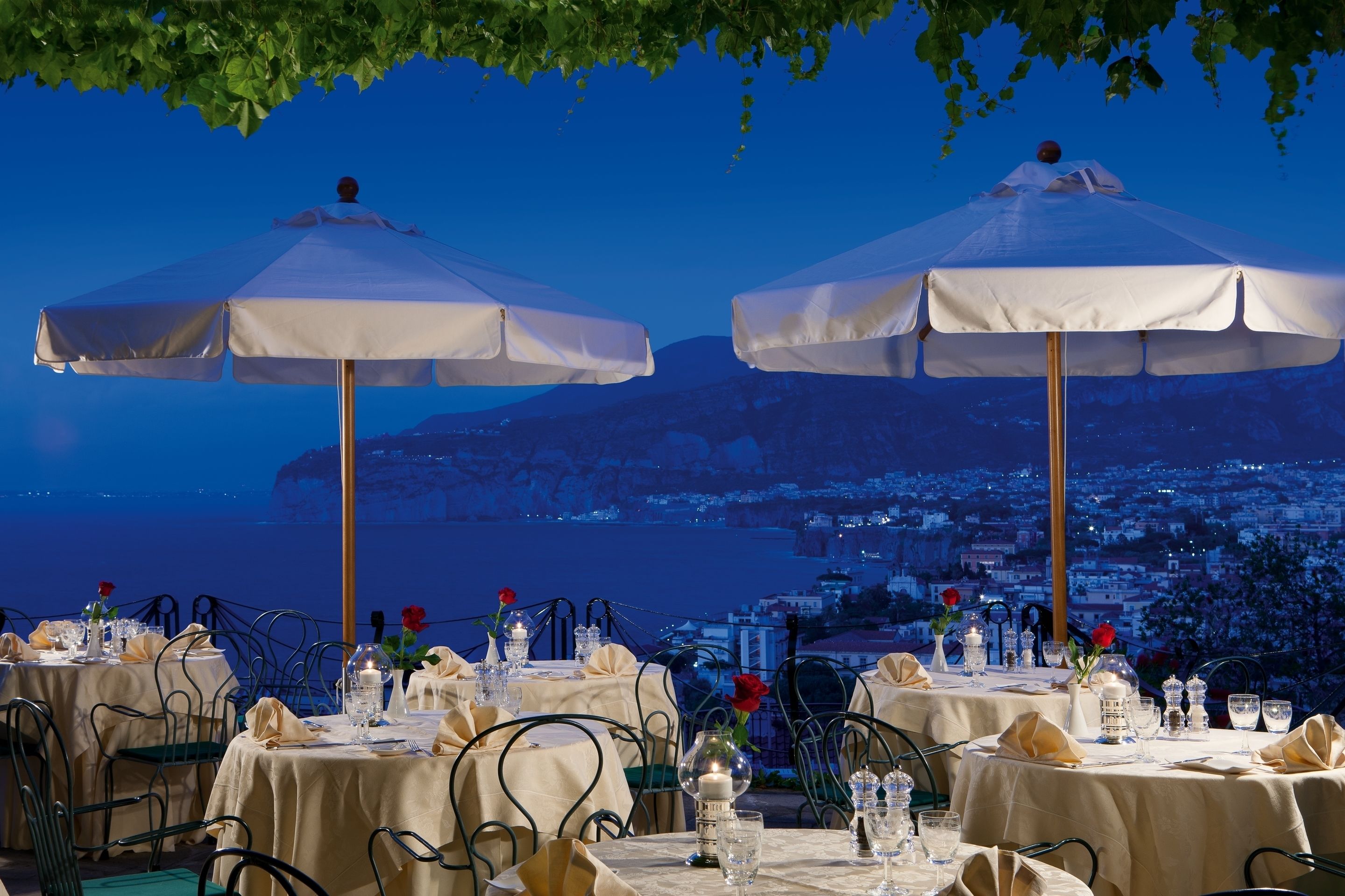 A restaurant with tables set for dining, white umbrellas, and a view of a coastal city at dusk.