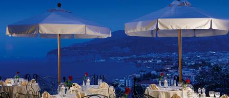 Un restaurante con mesas dispuestas para comer, sombrillas blancas y vistas a una ciudad costera al atardecer.