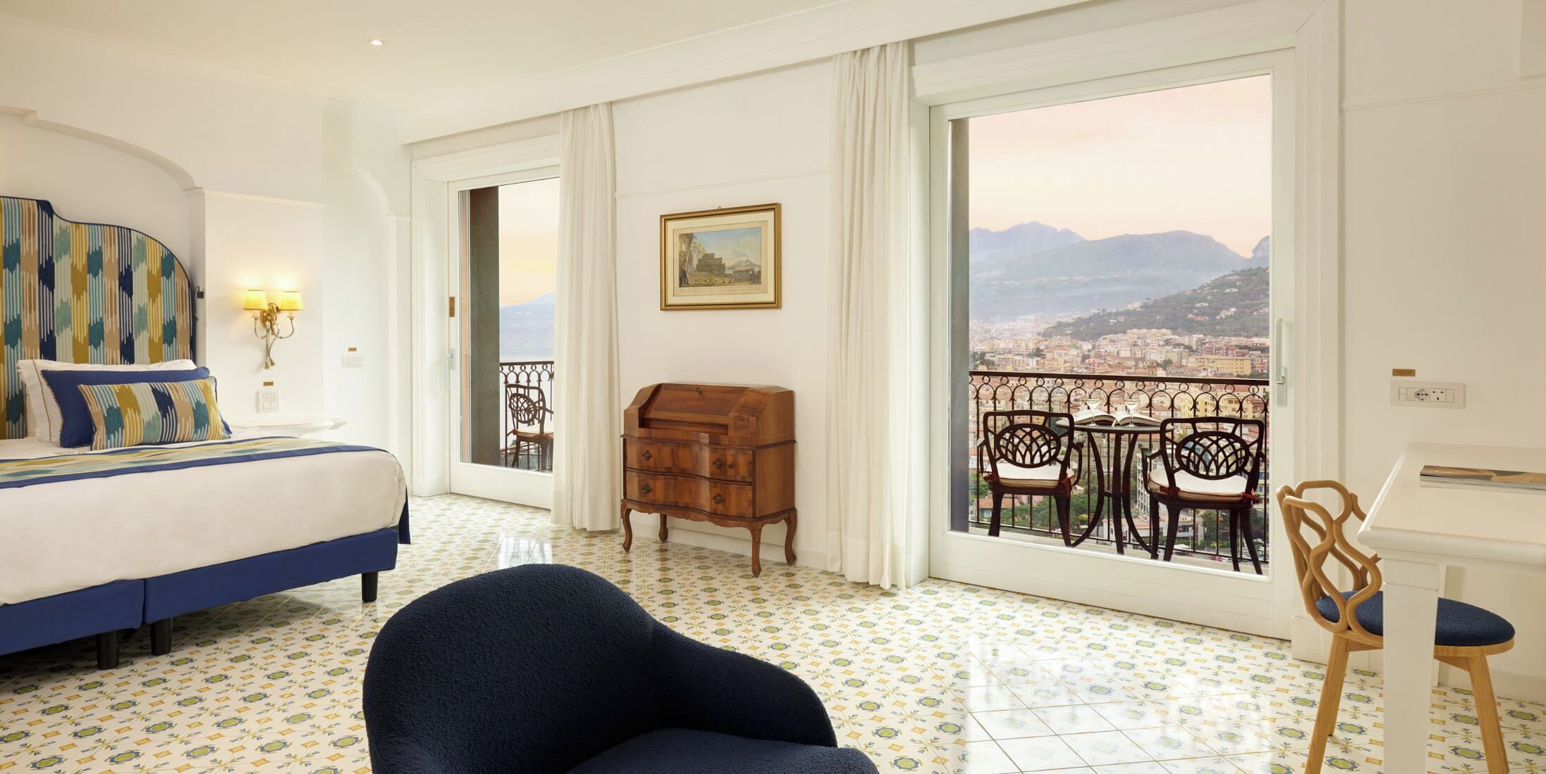 A hotel room with a bed, a chair, a wooden chest of drawers, and a balcony with a view of the city and mountains.