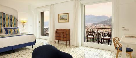 Habitación de hotel con cama, una silla, cómoda de madera y balcón con vistas a la ciudad y a las montañas.