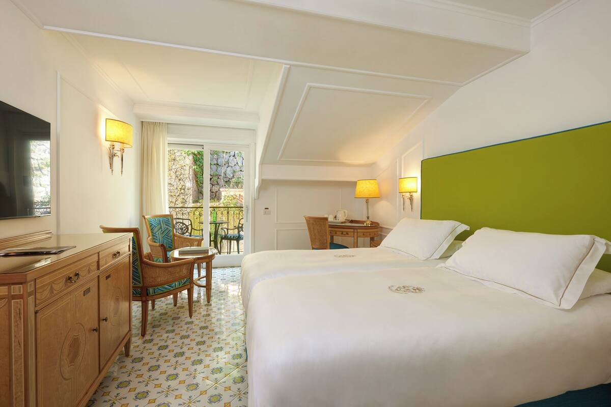 A hotel room with a large bed, a desk with a chair, a television, and a balcony with a view of greenery.