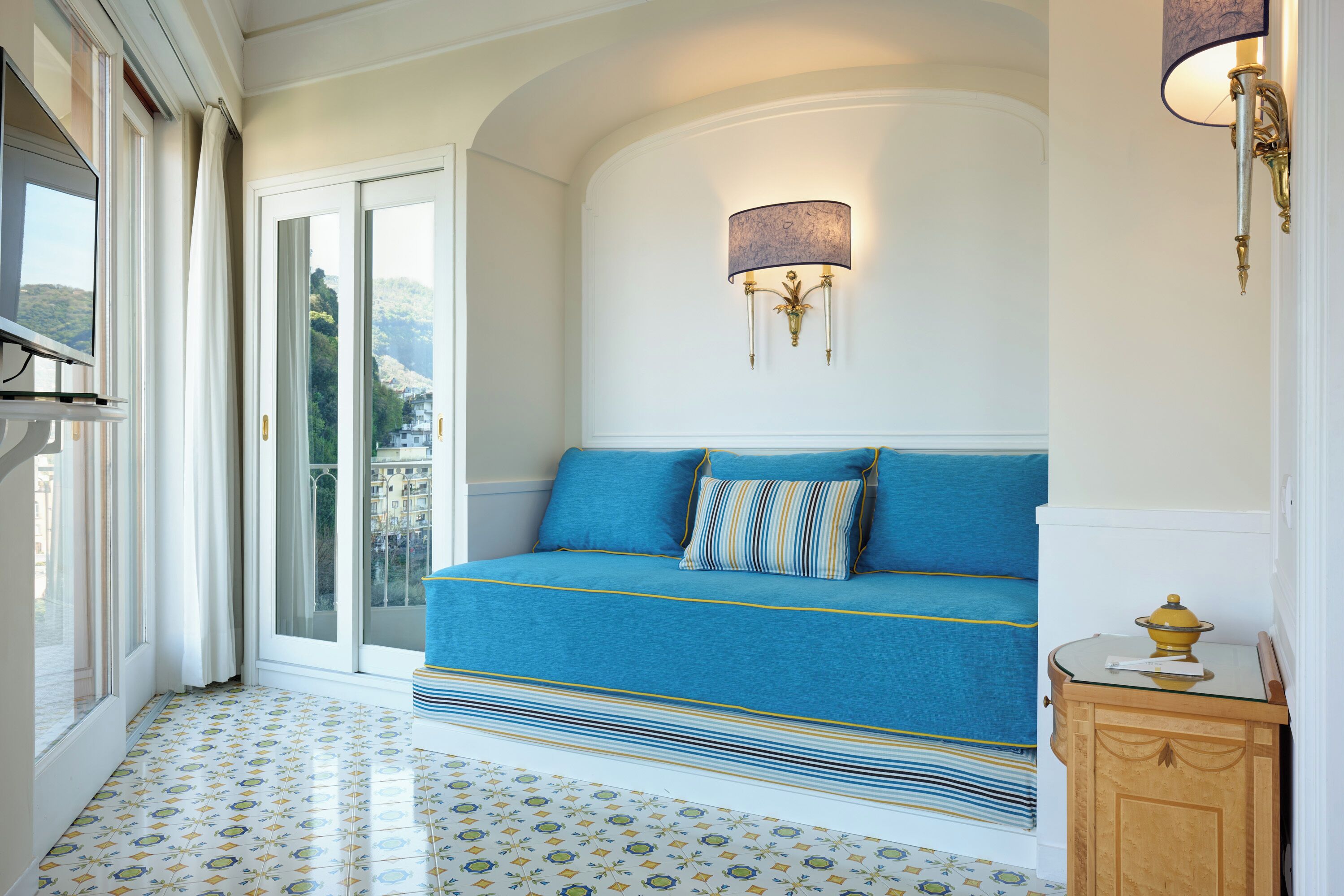 Quarto com sofá-cama, luminária de parede, pequeno armário e vista através de uma porta de vidro.