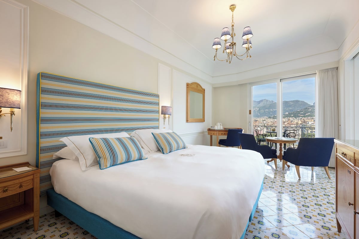 A hotel room with a large bed, a desk, chairs, a view of the city, and a chandelier.