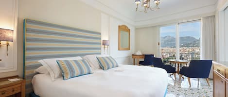A hotel room with a large bed, a desk, chairs, a view of the city, and a chandelier.