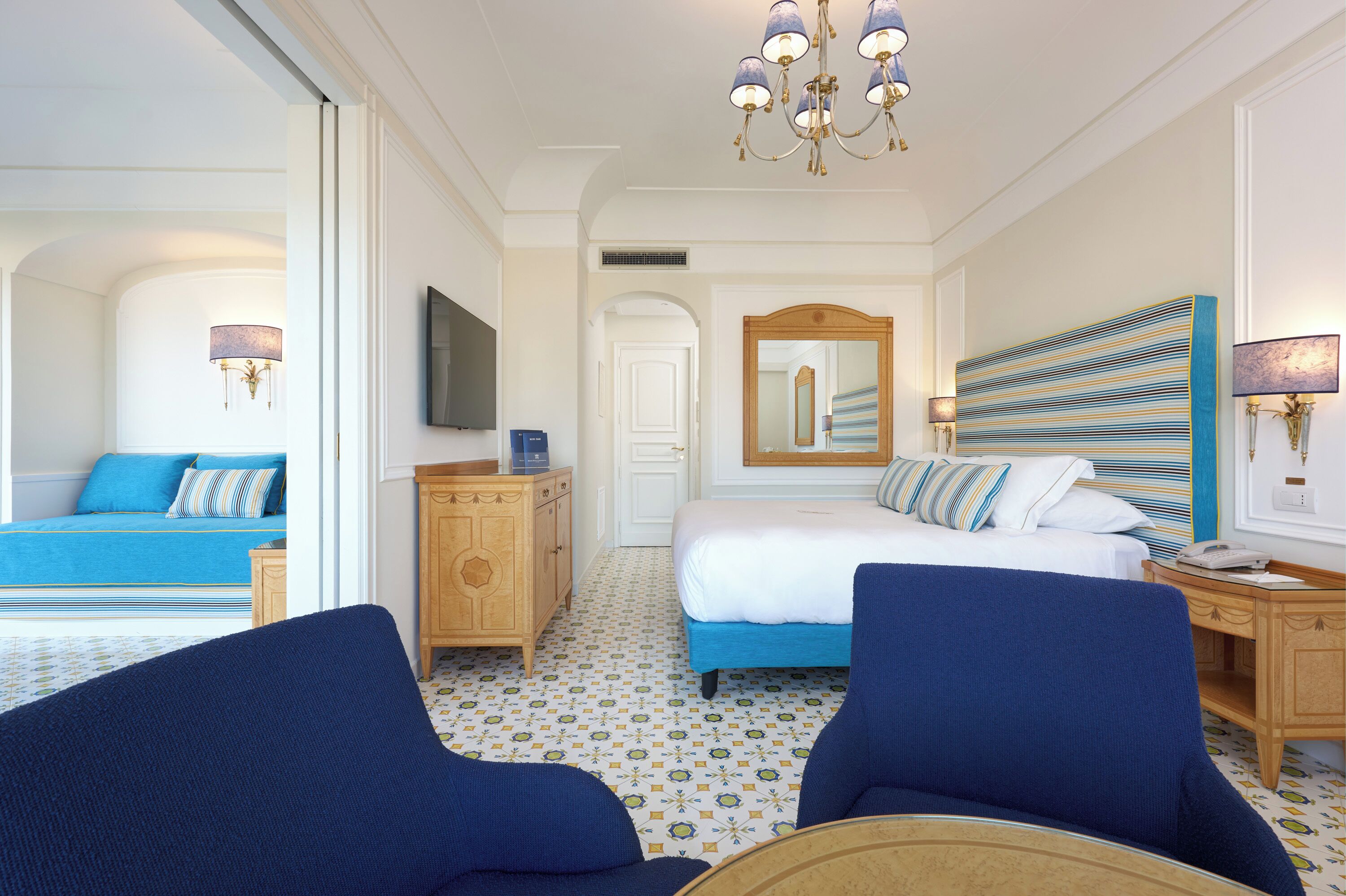 A hotel room with a bed, two blue armchairs, a wooden nightstand, a TV, and a chandelier.