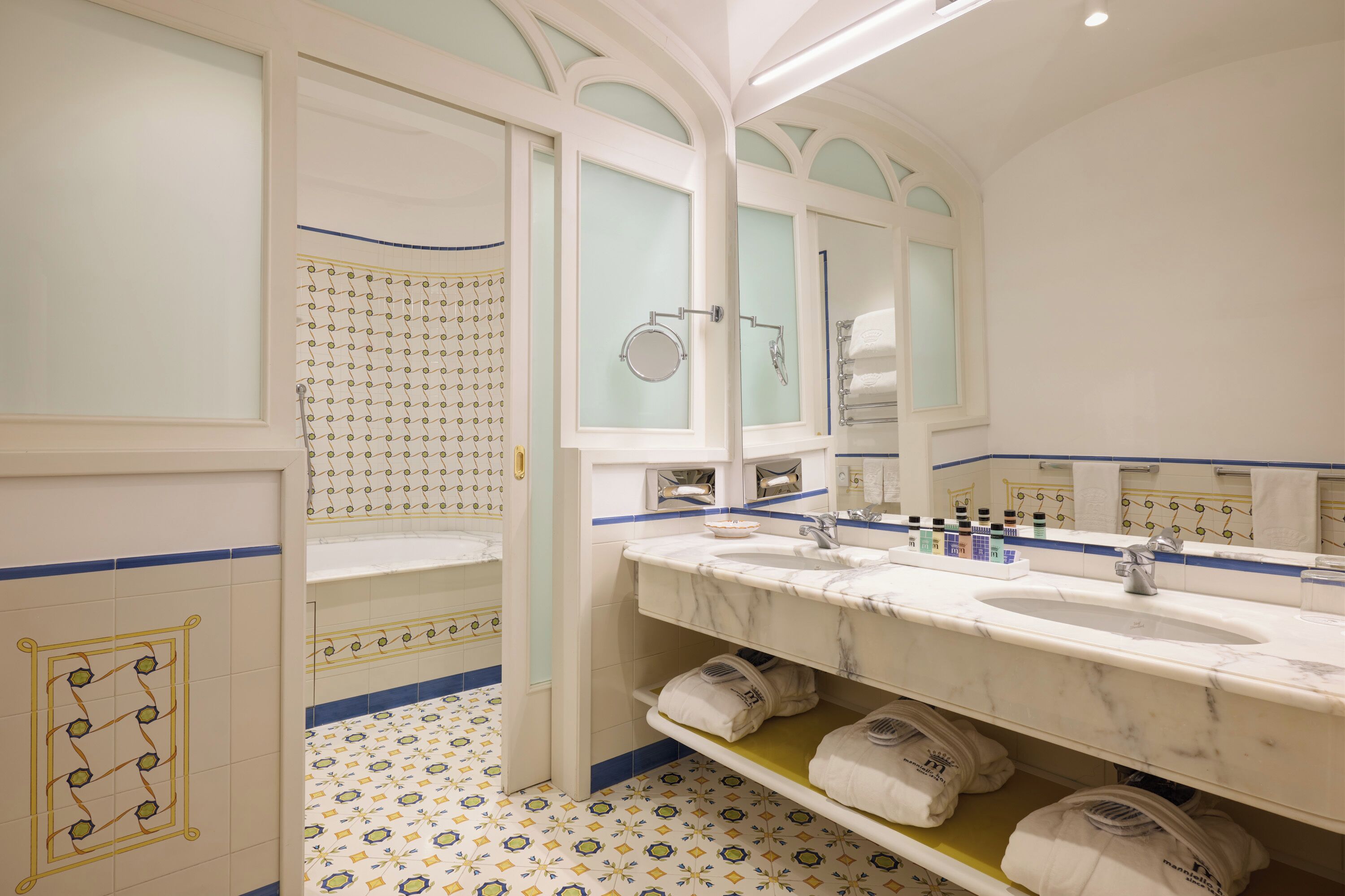 A bathroom with a large mirror, two sinks, a shower, and patterned floor tiles.