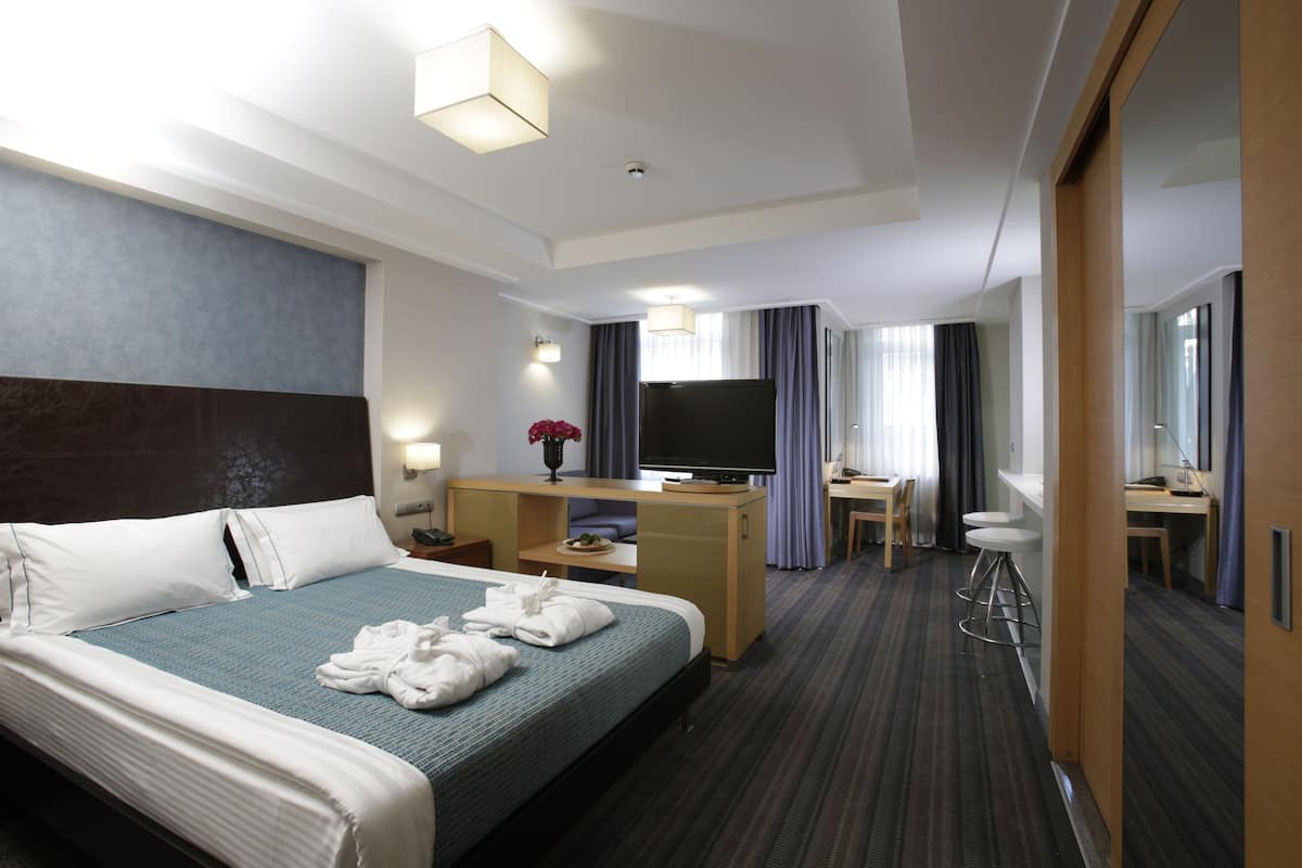 A hotel room with a bed, a desk with a TV, a chair, a small table, and a mirror.