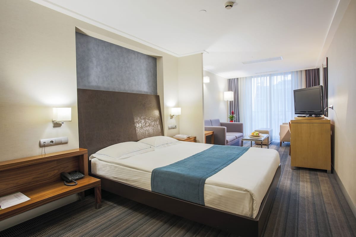 A hotel room with a large bed, bedside tables, a desk, a chair, a television, and a window with curtains.