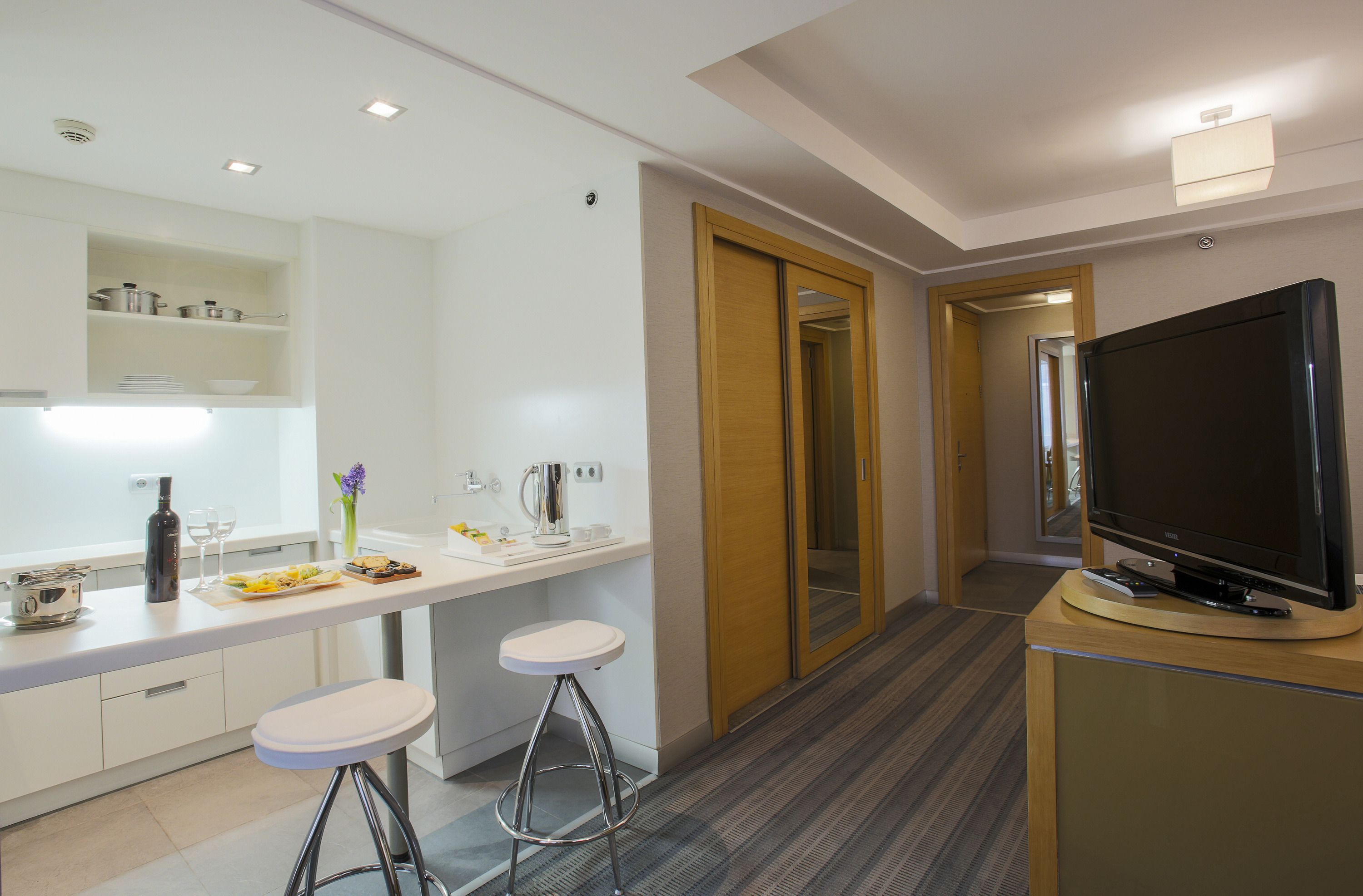 Una cocina moderna con encimera blanca, dos taburetes de bar, un televisor sobre un mueble de madera y una puerta de madera que da a otra habitación.