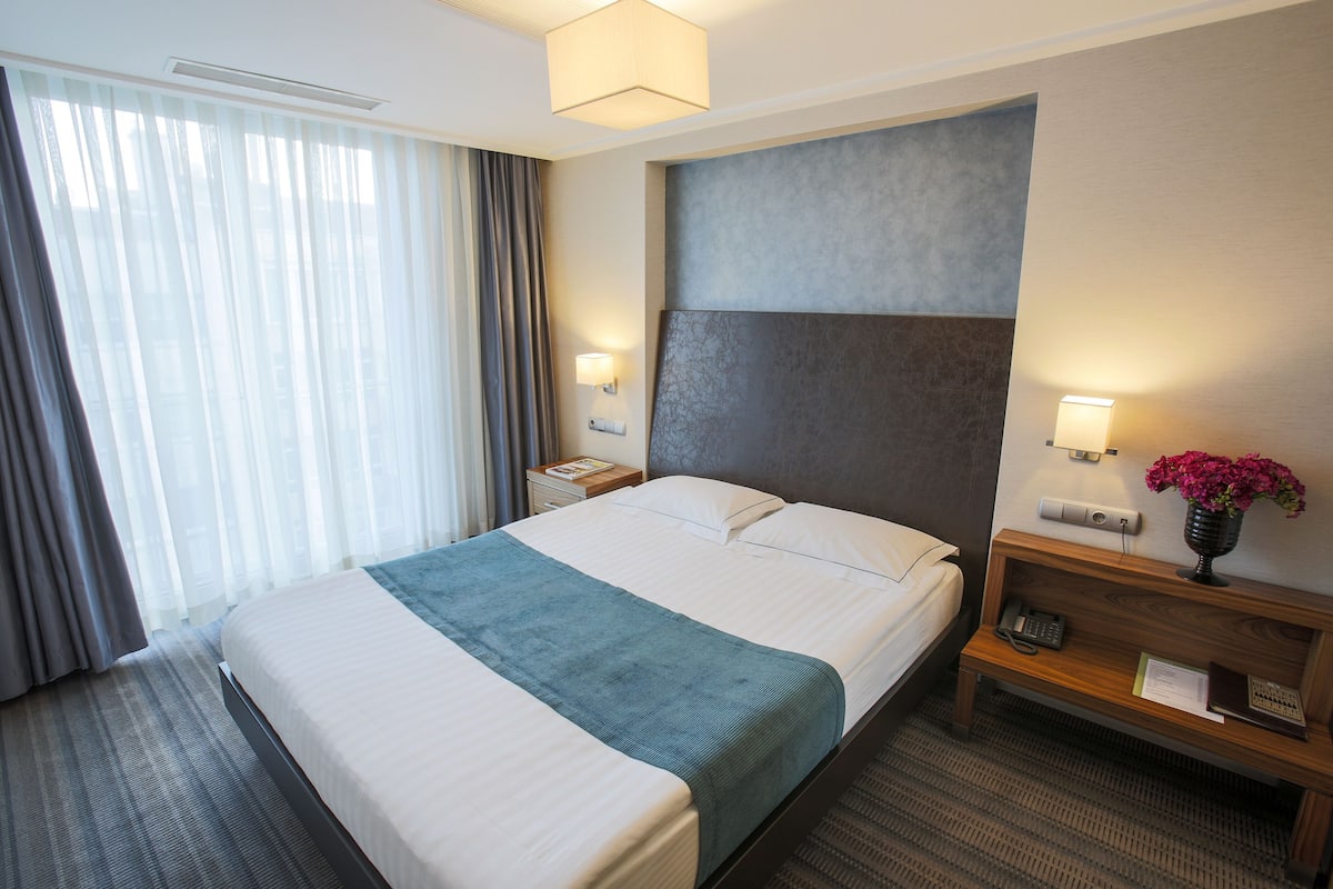 A hotel room with a large bed, a bedside table with a lamp, a nightstand with a phone, and a wall-mounted shelf with a vase of flowers.