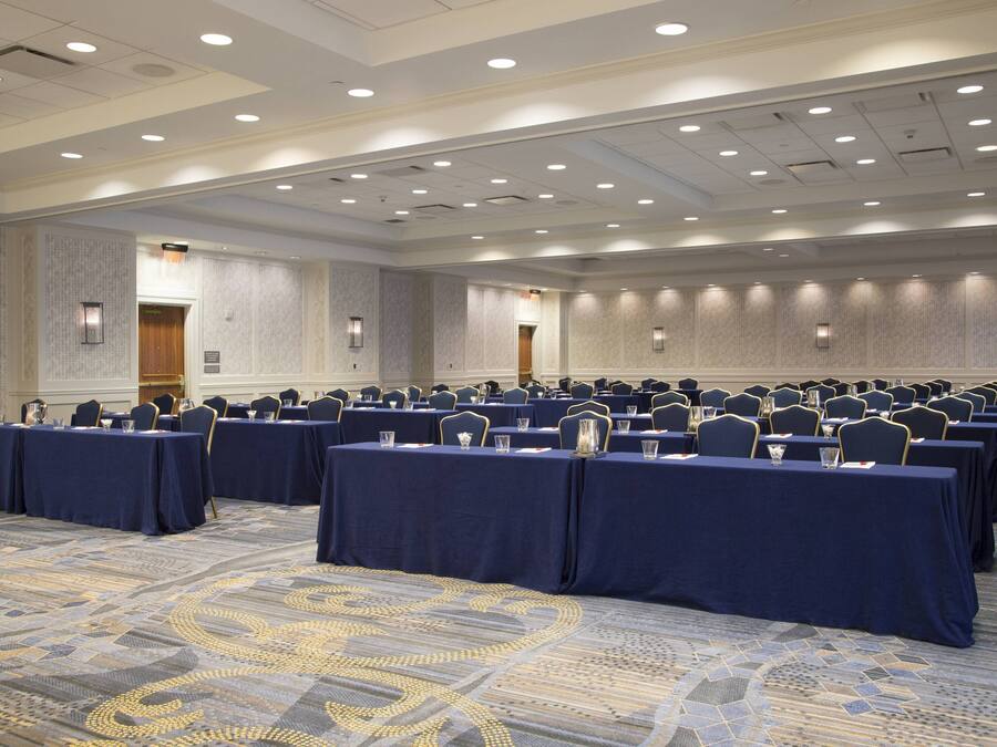 Un salón de conferencias con filas de mesas y sillas, piso alfombrado e iluminación empotrada.