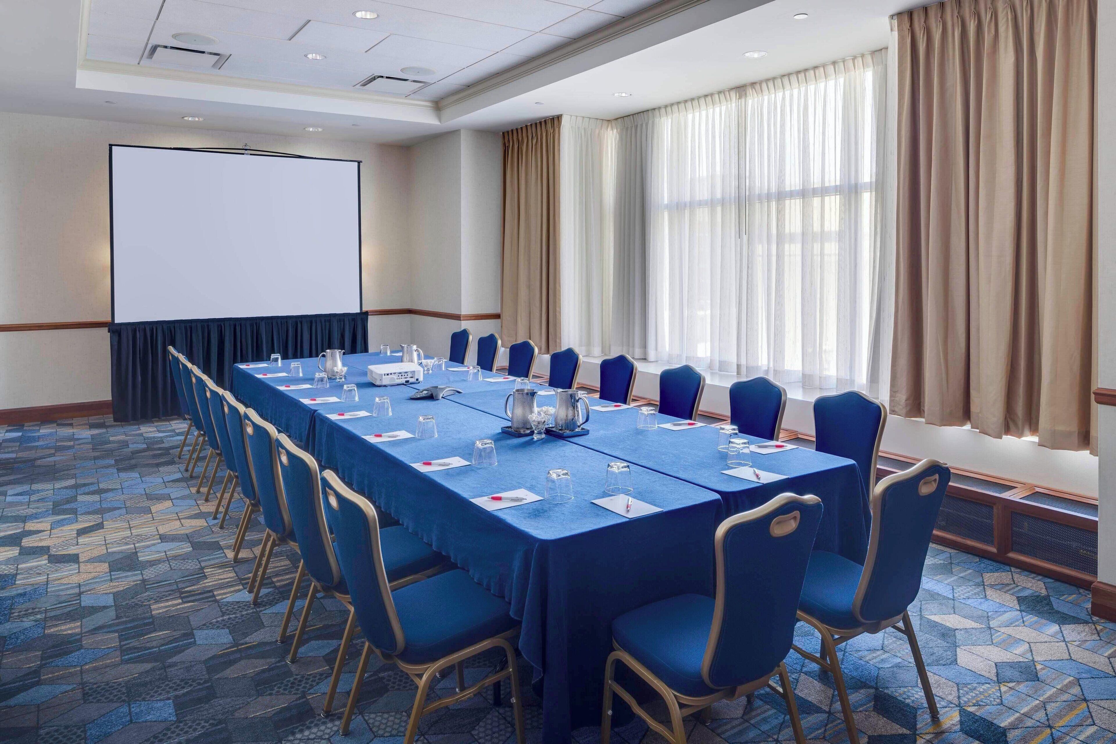 Una sala de conferencias con una larga mesa, sillas azules, una pantalla de proyector y grandes ventanales con cortinas.