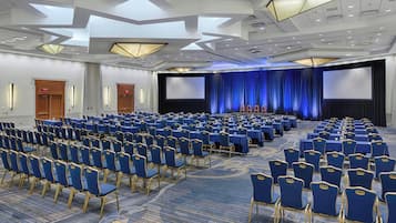 A large conference room with rows of blue chairs and tables, a stage with a blue curtain backdrop, and a large screen.