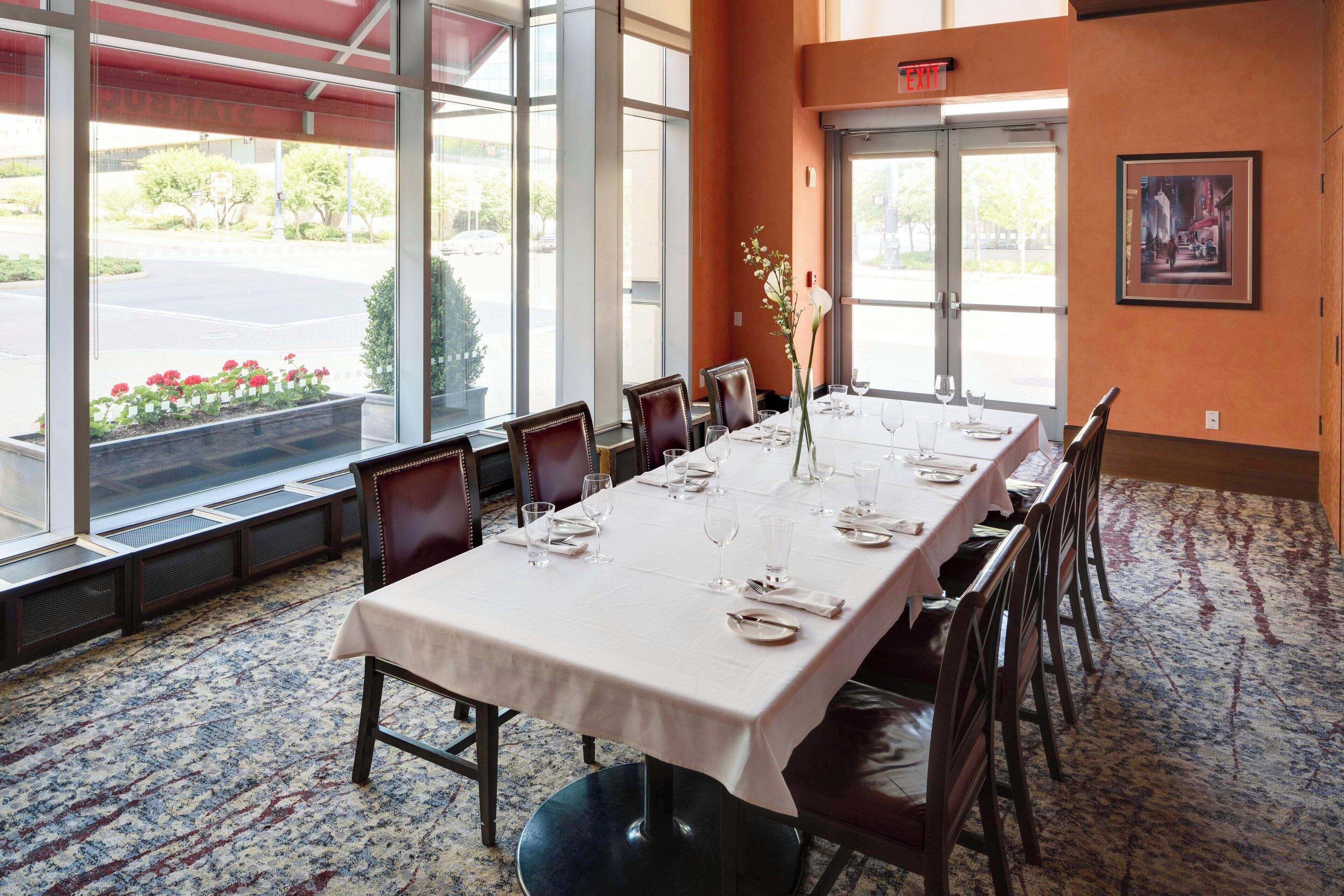A dining area with a long table set for six, large windows, and a view of the street outside.