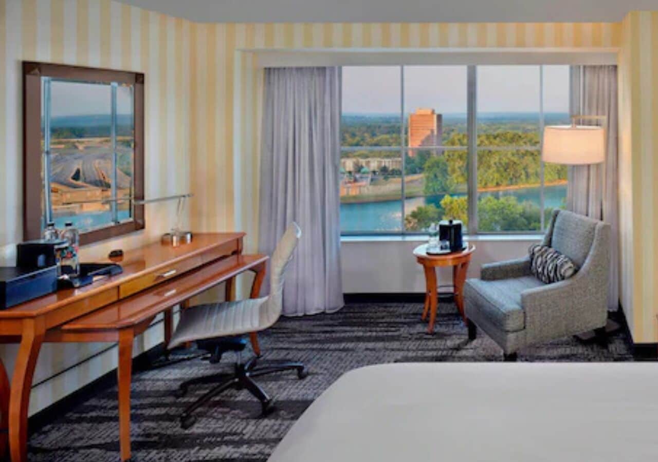 Quarto de hotel com uma cama grande, uma escrivaninha, uma cadeira, uma mesinha e vista para a paisagem pela janela.