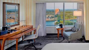 Quarto de hotel com uma cama grande, uma escrivaninha, uma cadeira, uma mesinha e vista para a paisagem pela janela.