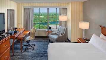 A hotel room with a large bed, a desk with a computer, a chair, a TV, and a view of a lake and greenery.