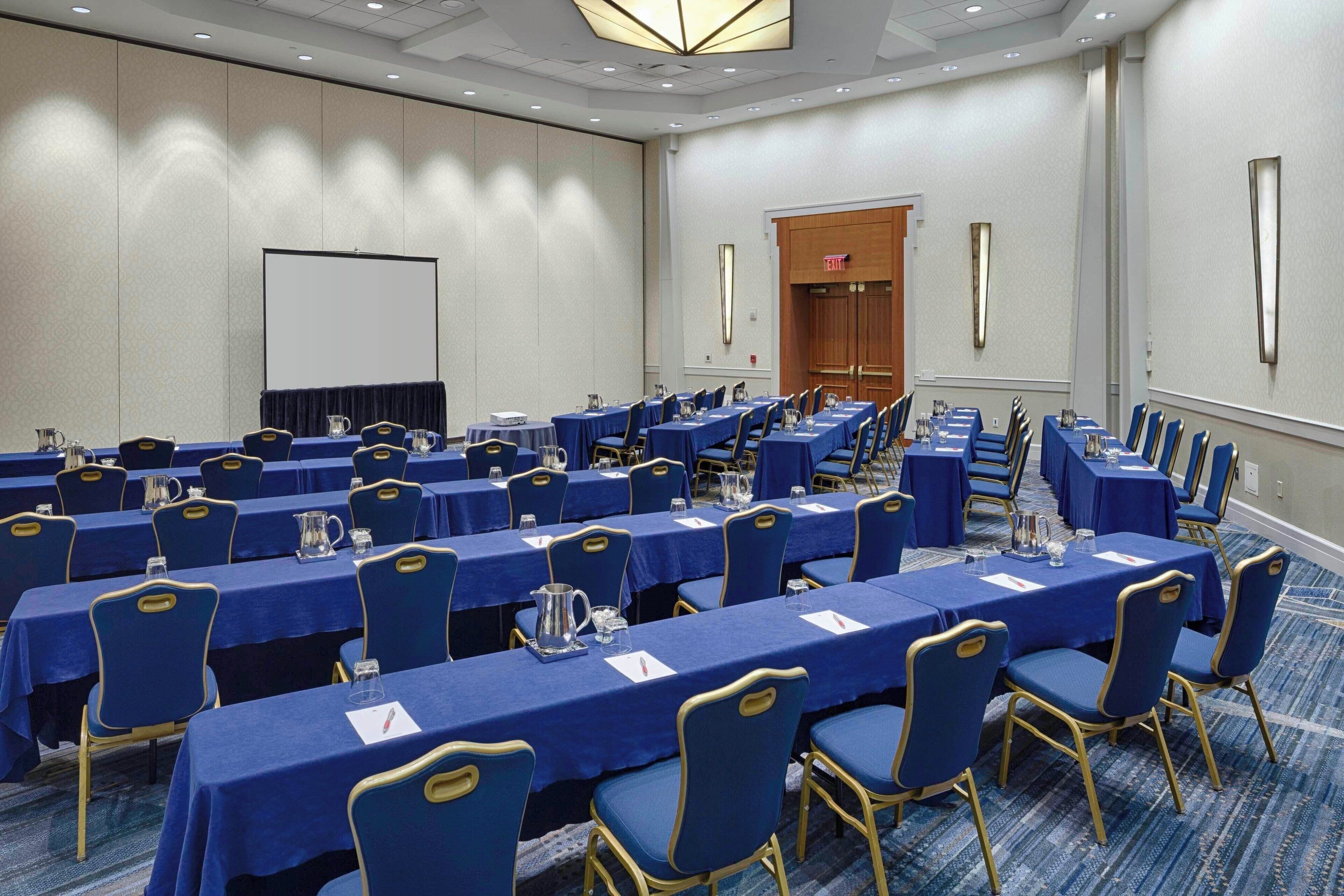 A conference room with multiple tables and chairs, a projector screen, and a door with a sign.