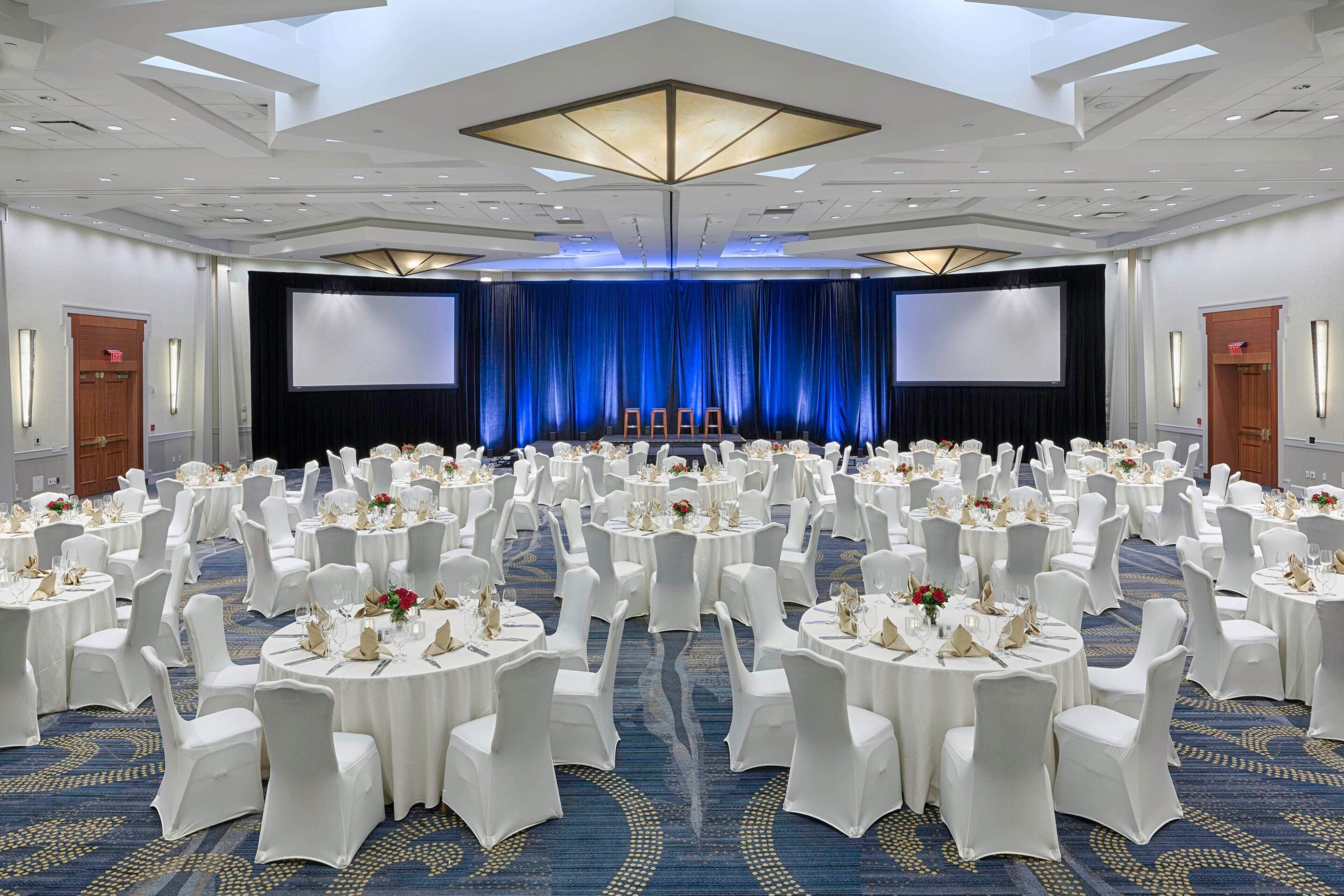 A large conference room with round tables and white chairs, a stage with a blue backdrop, and a high ceiling with recessed lighting.