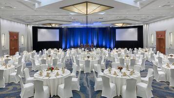 A large conference room with round tables and white chairs, a stage with a blue backdrop, and a high ceiling with recessed lighting.