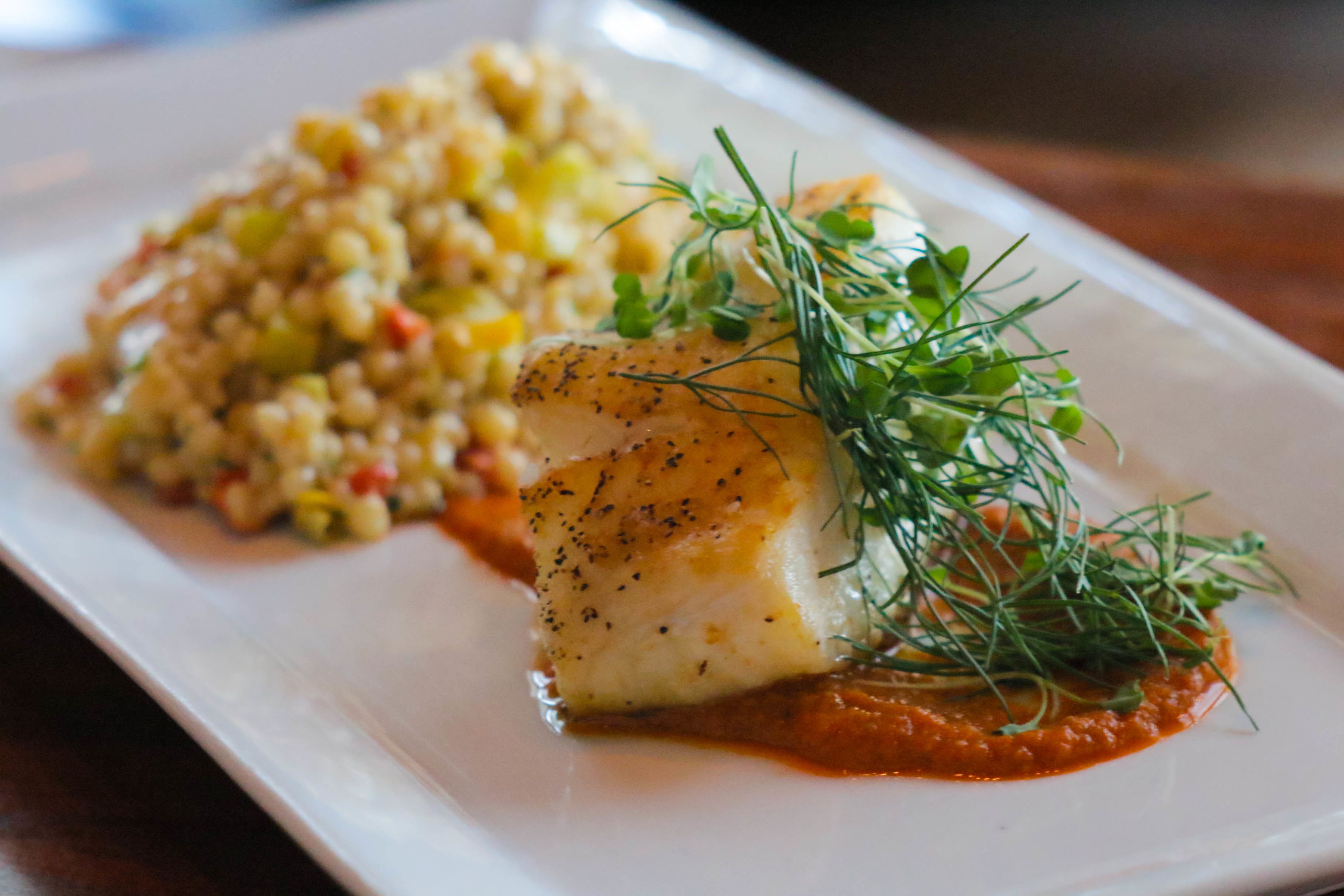En hvid, rektangulær tallerken med et stykke fisk, drysset med dild og sort peber, samt en portion couscous.