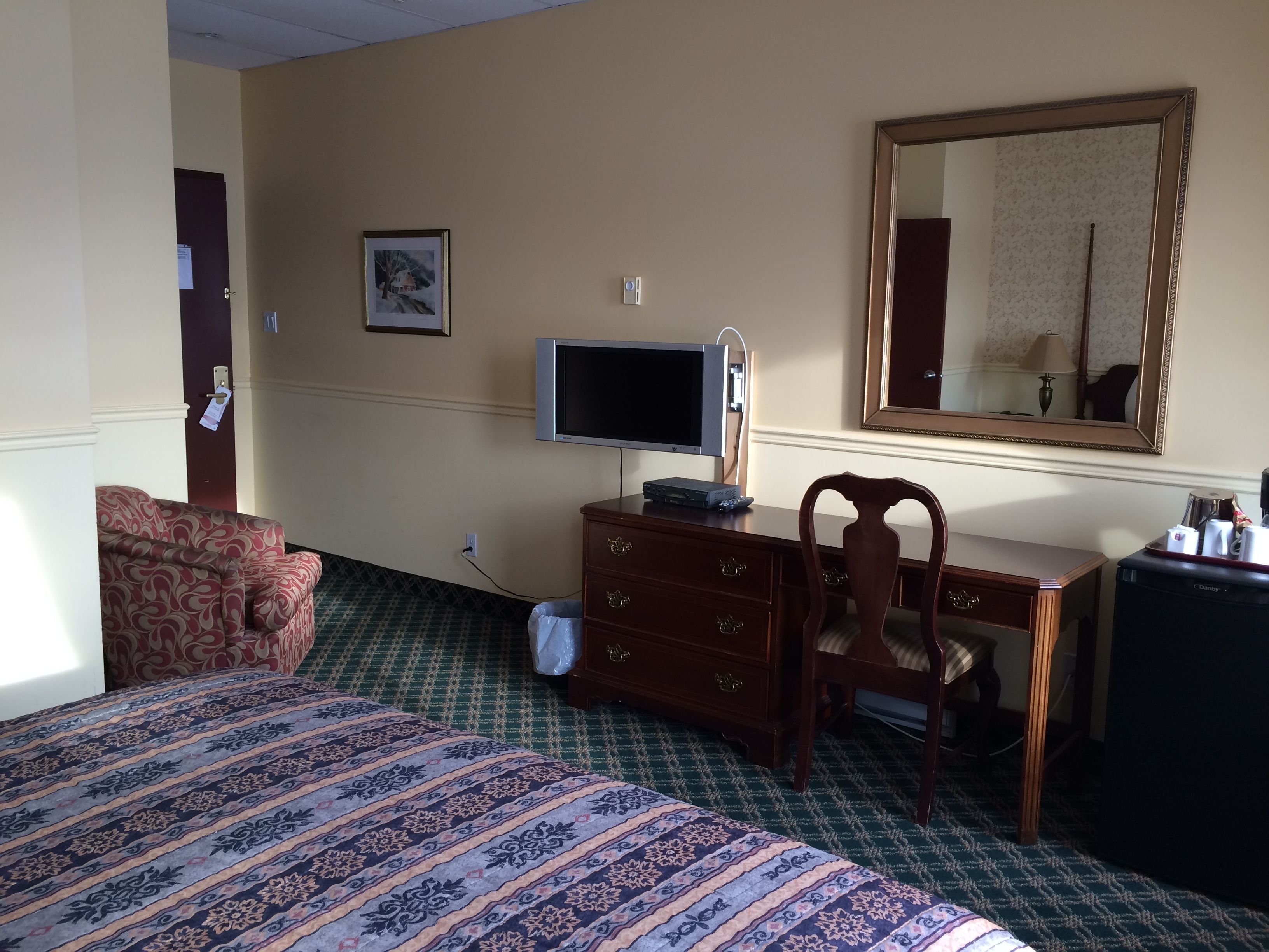 Quarto de hotel com cama, escrivaninha com computador, cadeira, cômoda, espelho e televisão.