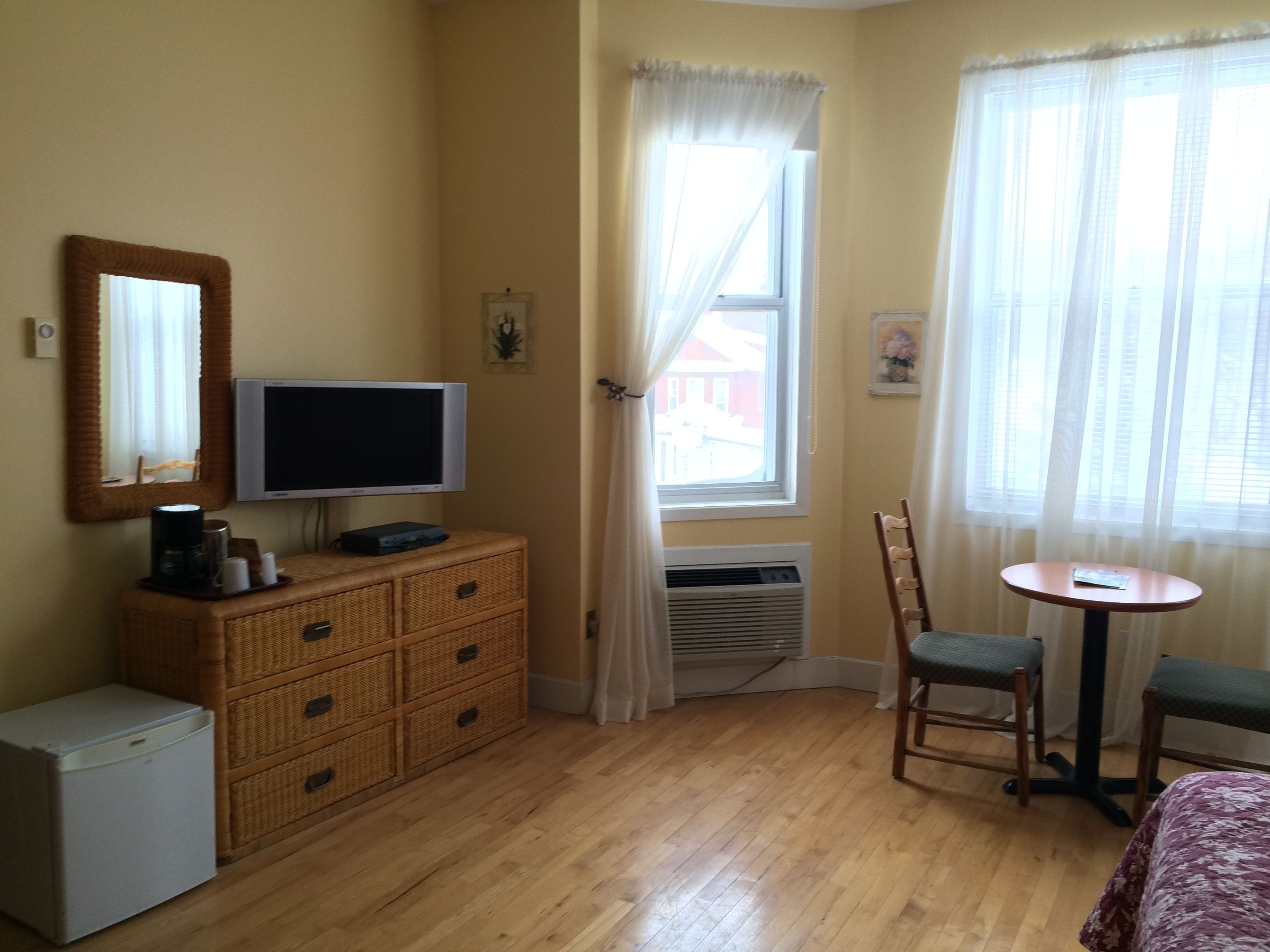 Une chambre avec un téléviseur posé sur une commode, un petit réfrigérateur, une chaise, une table et une fenêtre avec des rideaux.