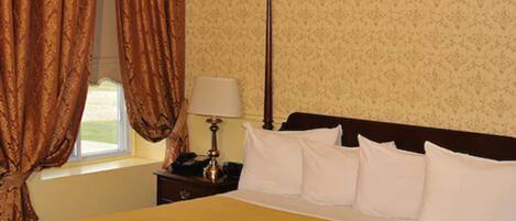 A neatly made bed with white pillows and a gold bedspread in a room with patterned wallpaper and a window with curtains.