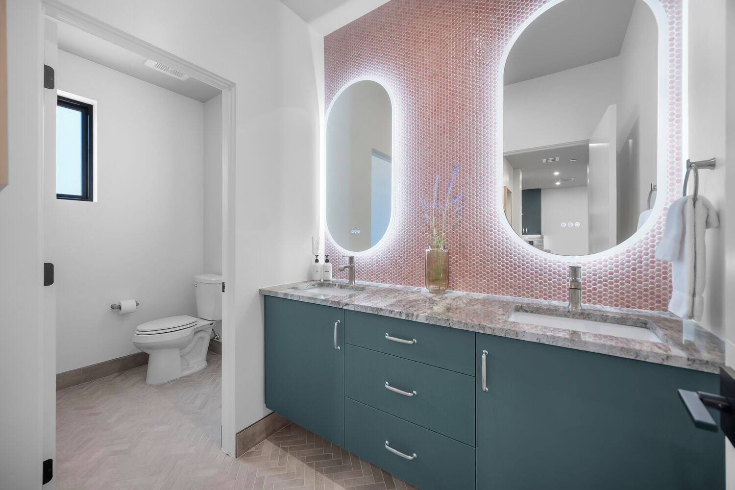 A modern bathroom with a teal vanity, a large mirror, and a pink patterned wall.