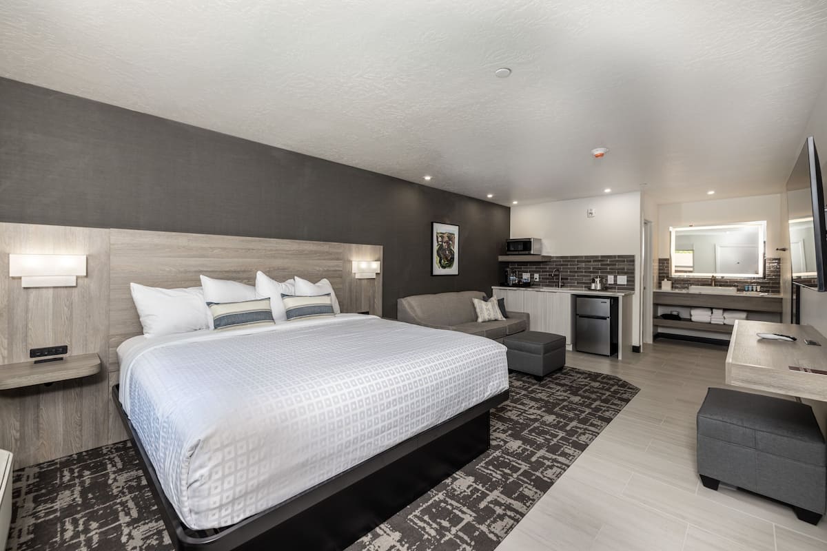 A modern hotel room with a large bed, a sofa, a small kitchenette, and a bathroom.