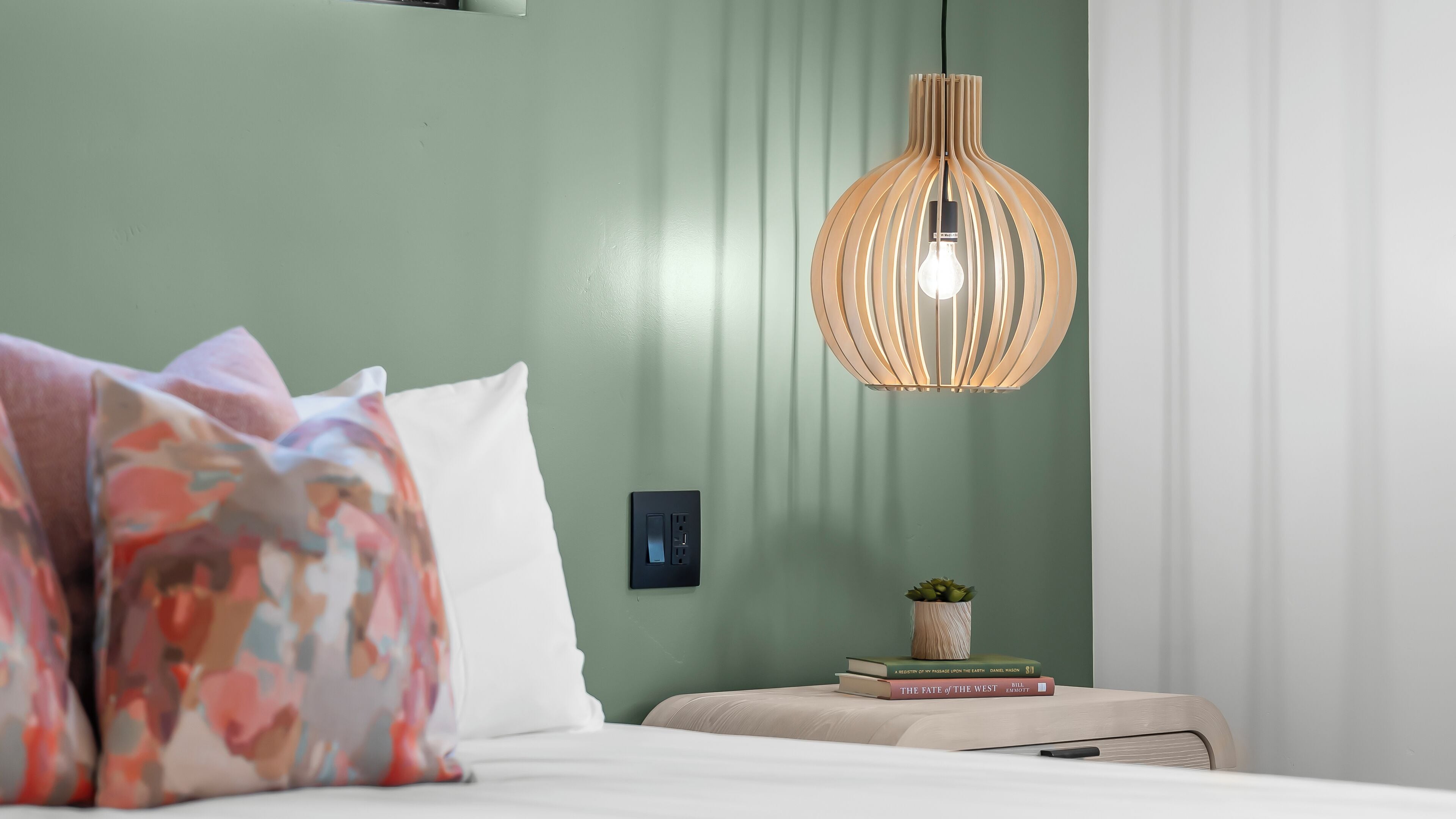 A neatly made bed with floral pillows, a bedside table with books and a small plant, and a modern pendant light.
