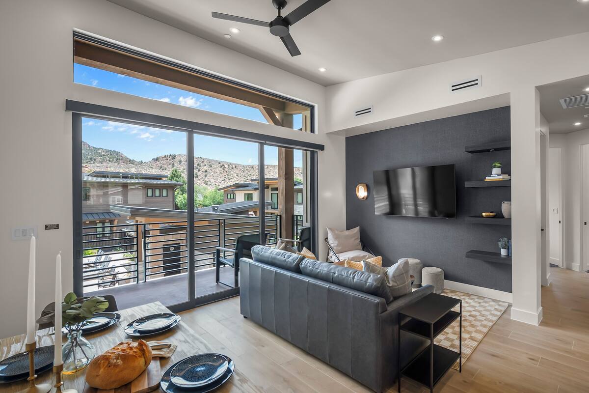 A spacious living room with a large sofa, a flat-screen TV, and a balcony with a view of the surrounding area.