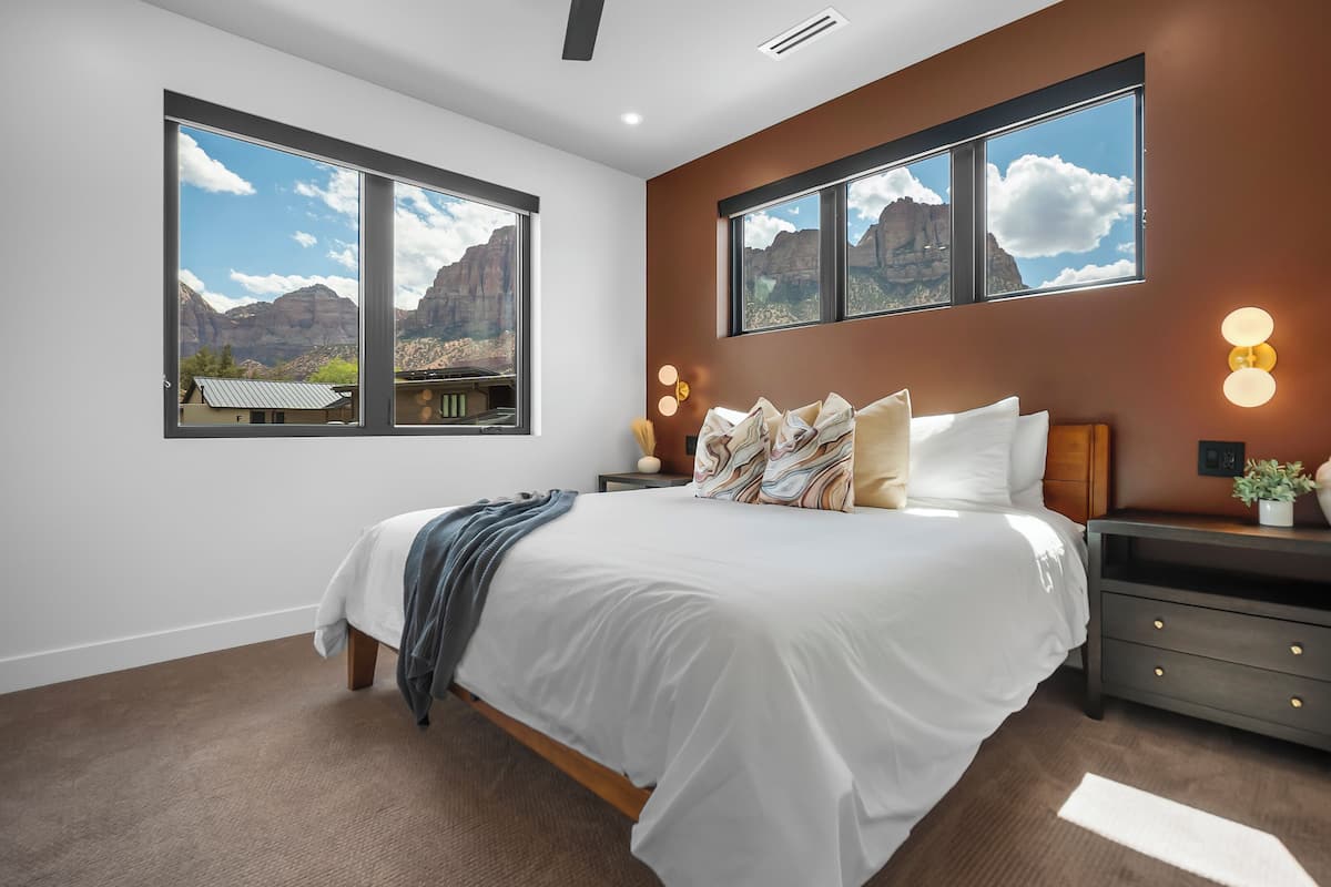 A bedroom with a large bed, two bedside tables, a nightstand, and a view of mountains through the windows.