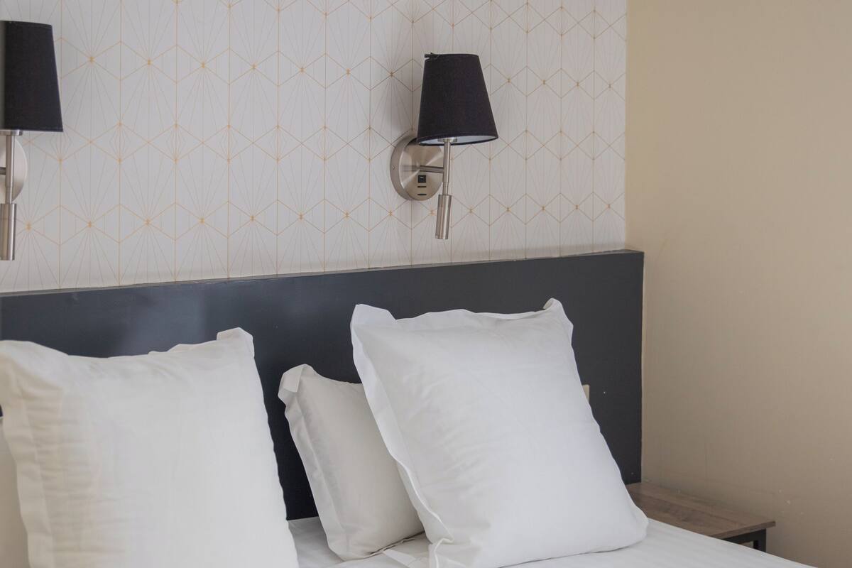Una cama bien hecha con sábanas y almohadas blancas, cabecera oscura y dos lámparas fijadas a la pared.