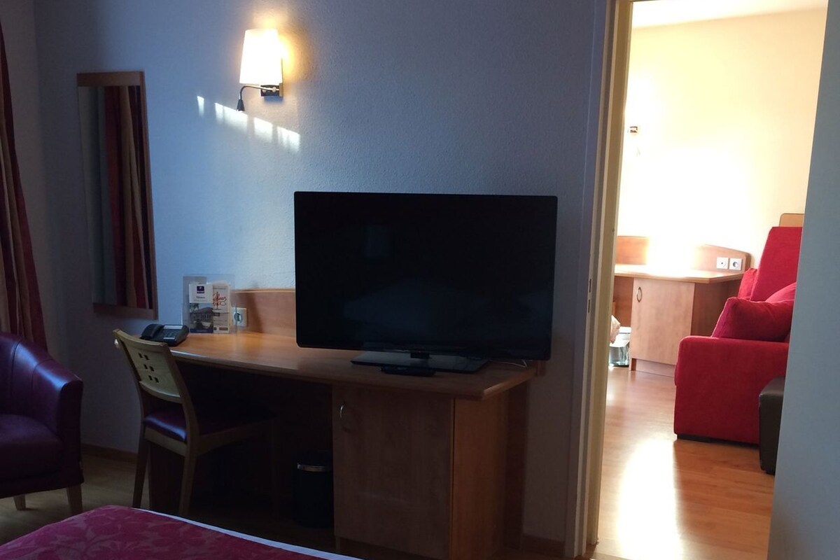 Una habitación de hotel con una cama, un escritorio con televisor, una silla, un sofá rojo y una puerta que da a otra habitación.