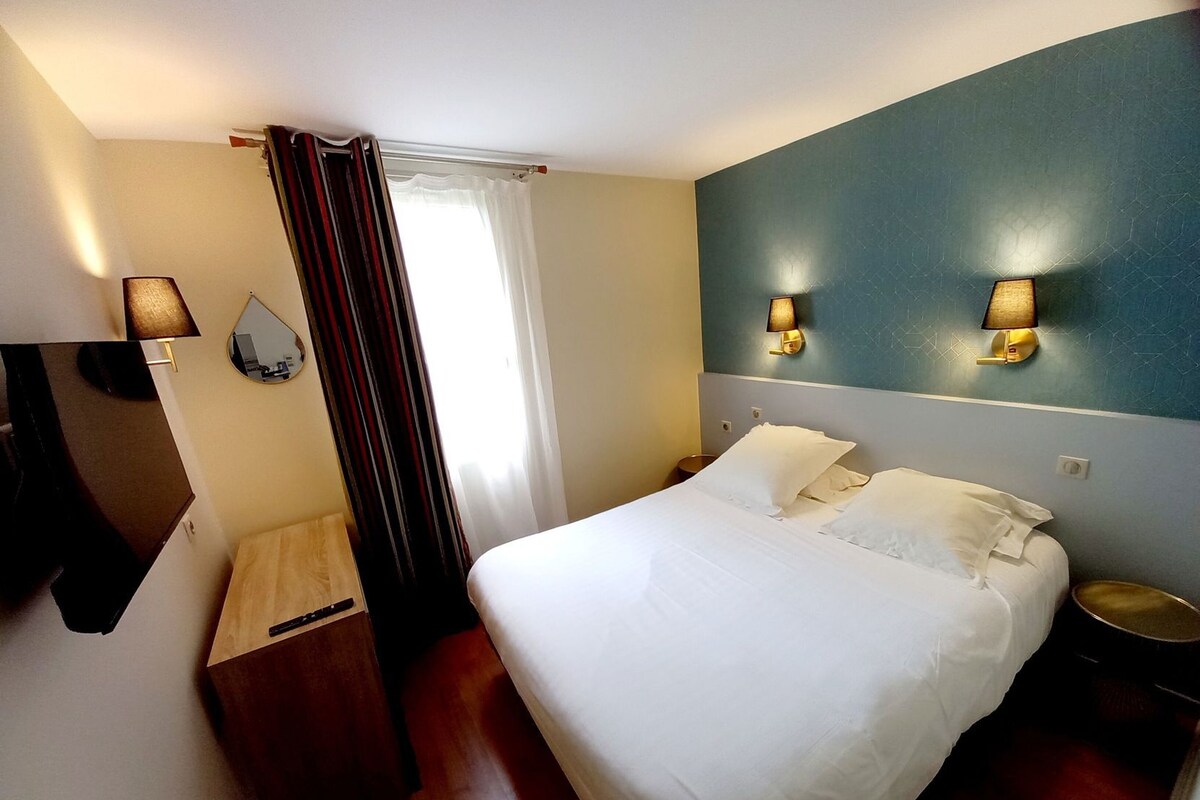 Una habitación de hotel con cama, mesita de noche, lámpara, espejo y ventana con cortinas.
