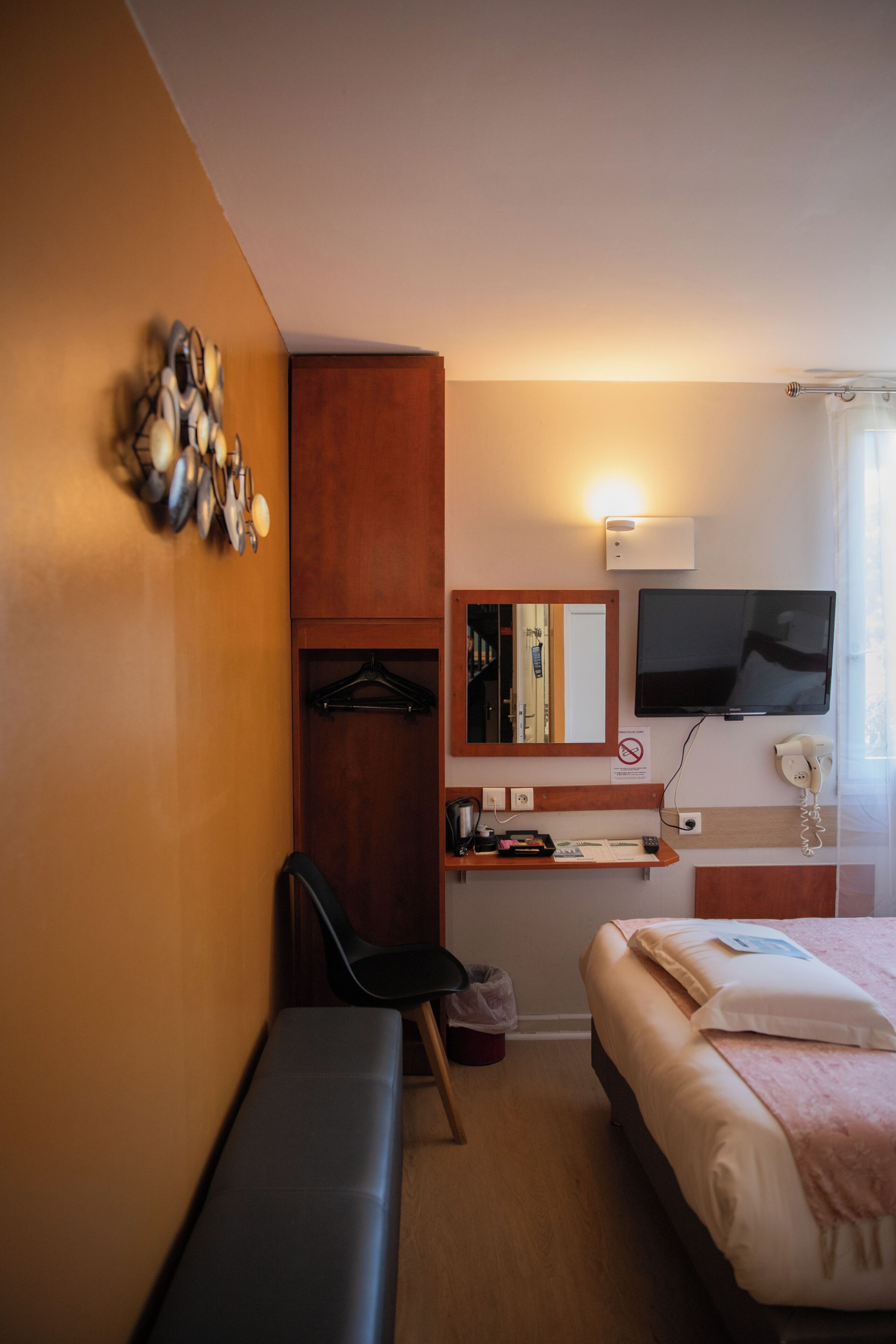 A hotel room with a bed, a bench, a TV, a coat rack, a mirror, and a wall-mounted telephone.