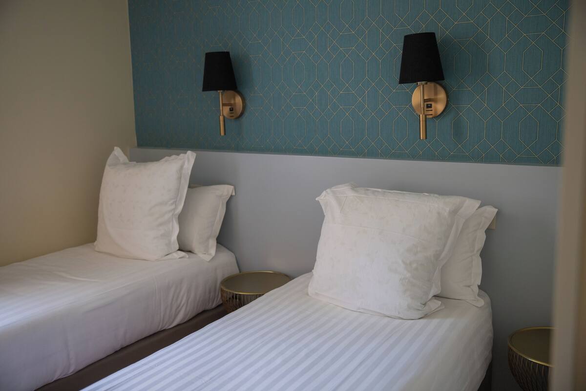Habitación de hotel con dos camas individuales, una mesita redonda, una lámpara de pared y un papel tapiz estampado.