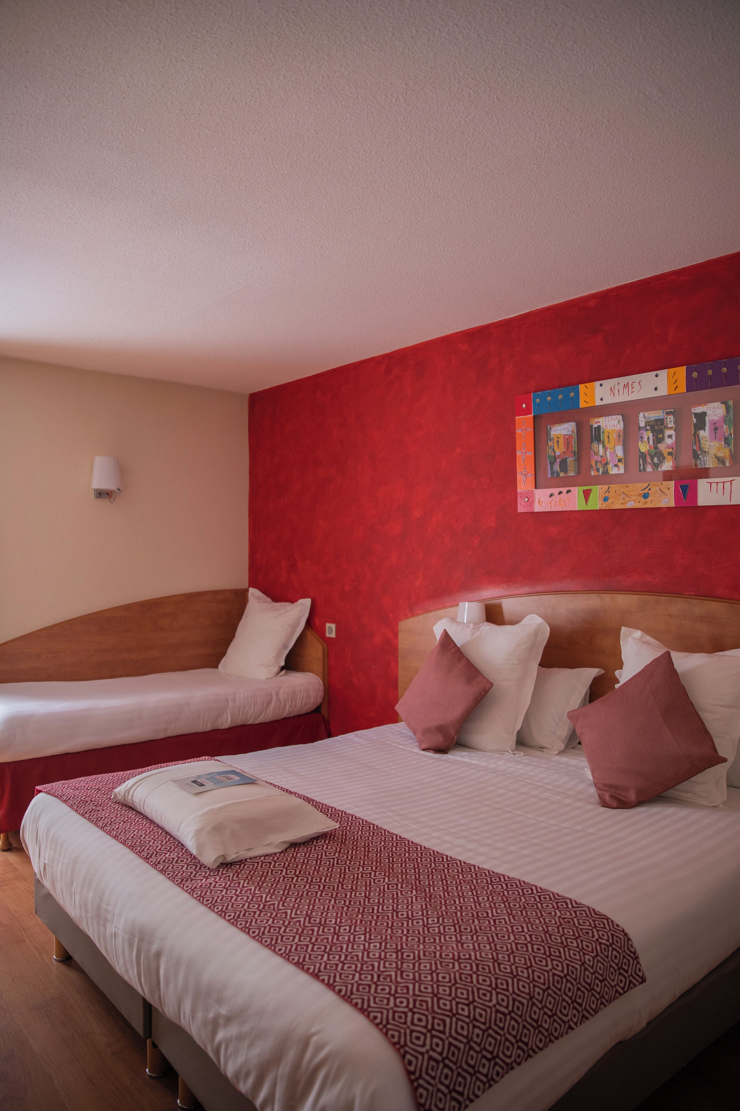 A hotel room with two beds, a red accent wall, a bulletin board with photos, and a white lamp.