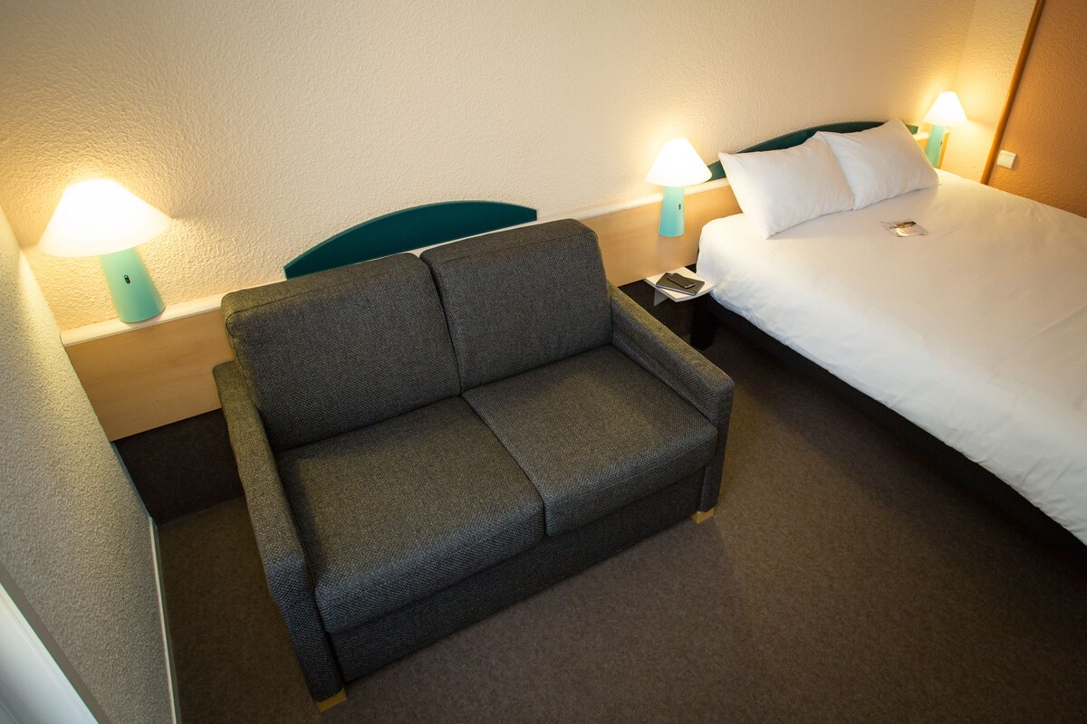 Een hotelkamer met een grijze bank, een bed met wit beddengoed en twee bedlampjes.