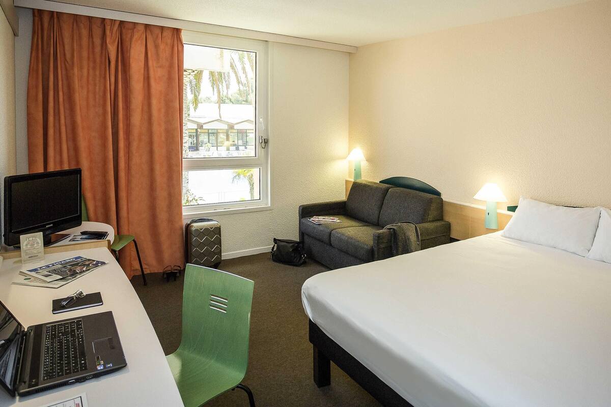 Een hotelkamer met een bed, een bureau met een laptop, een stoel, een televisie en een raam met gordijnen.
