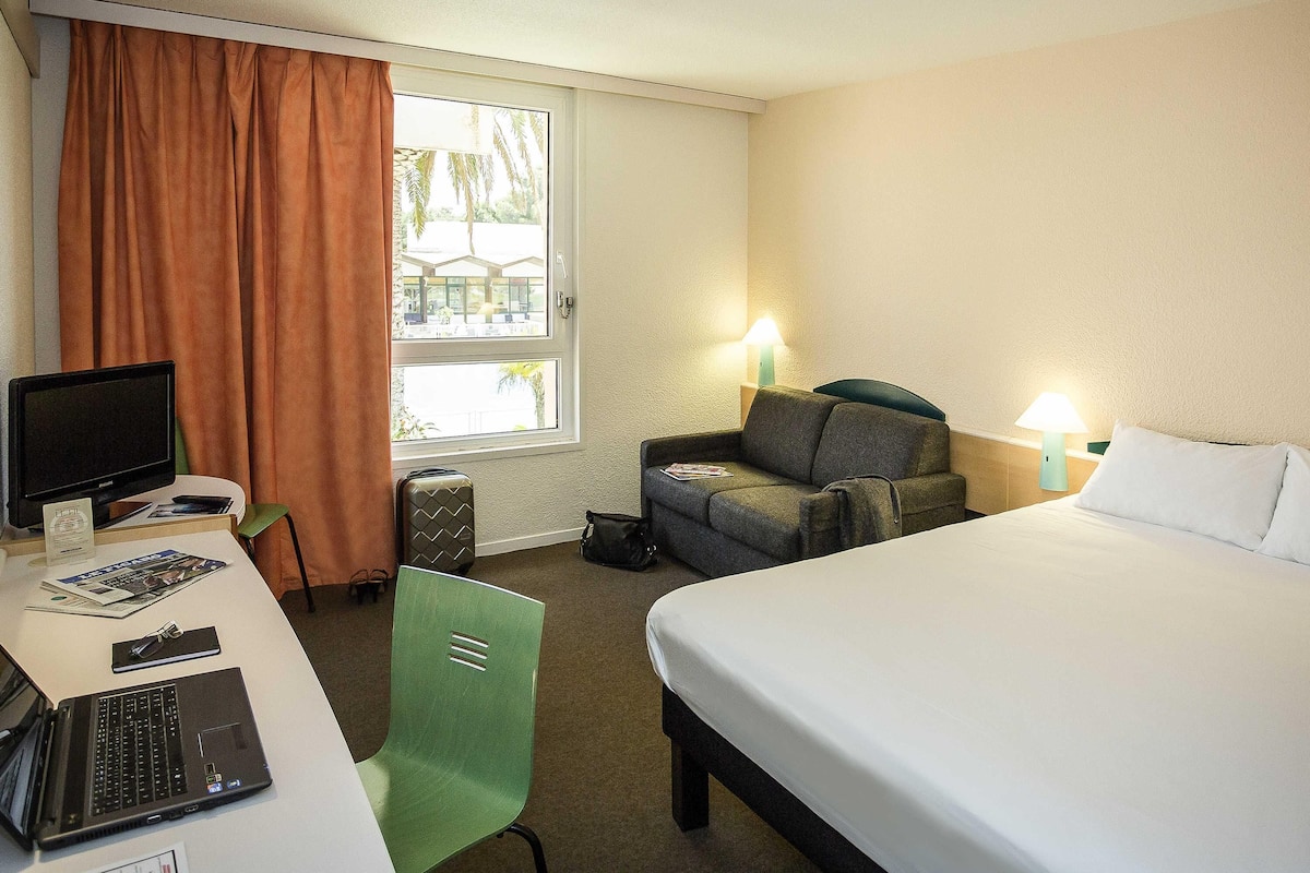 A hotel room with a bed, a desk with a laptop, a chair, a television, and a window with curtains.
