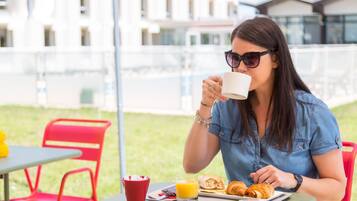 A person seated at an outdoor table with a meal, including croissants and orange juice, in a setting with red chairs and a building in the background.