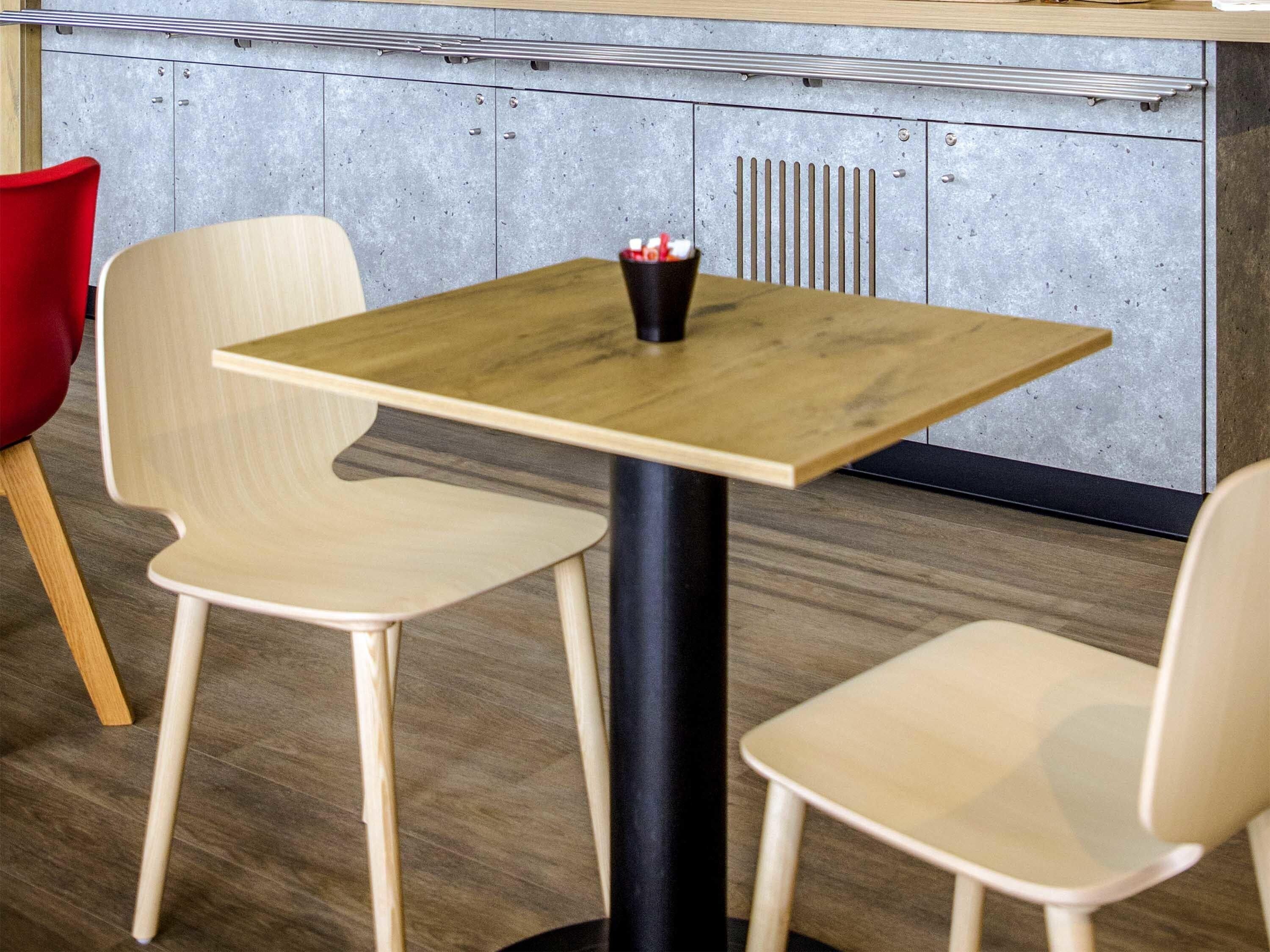 Ein kleiner Tisch mit schwarzem Gestell und Holzplatte, begleitet von zwei Stühlen mit heller Holzoptik.