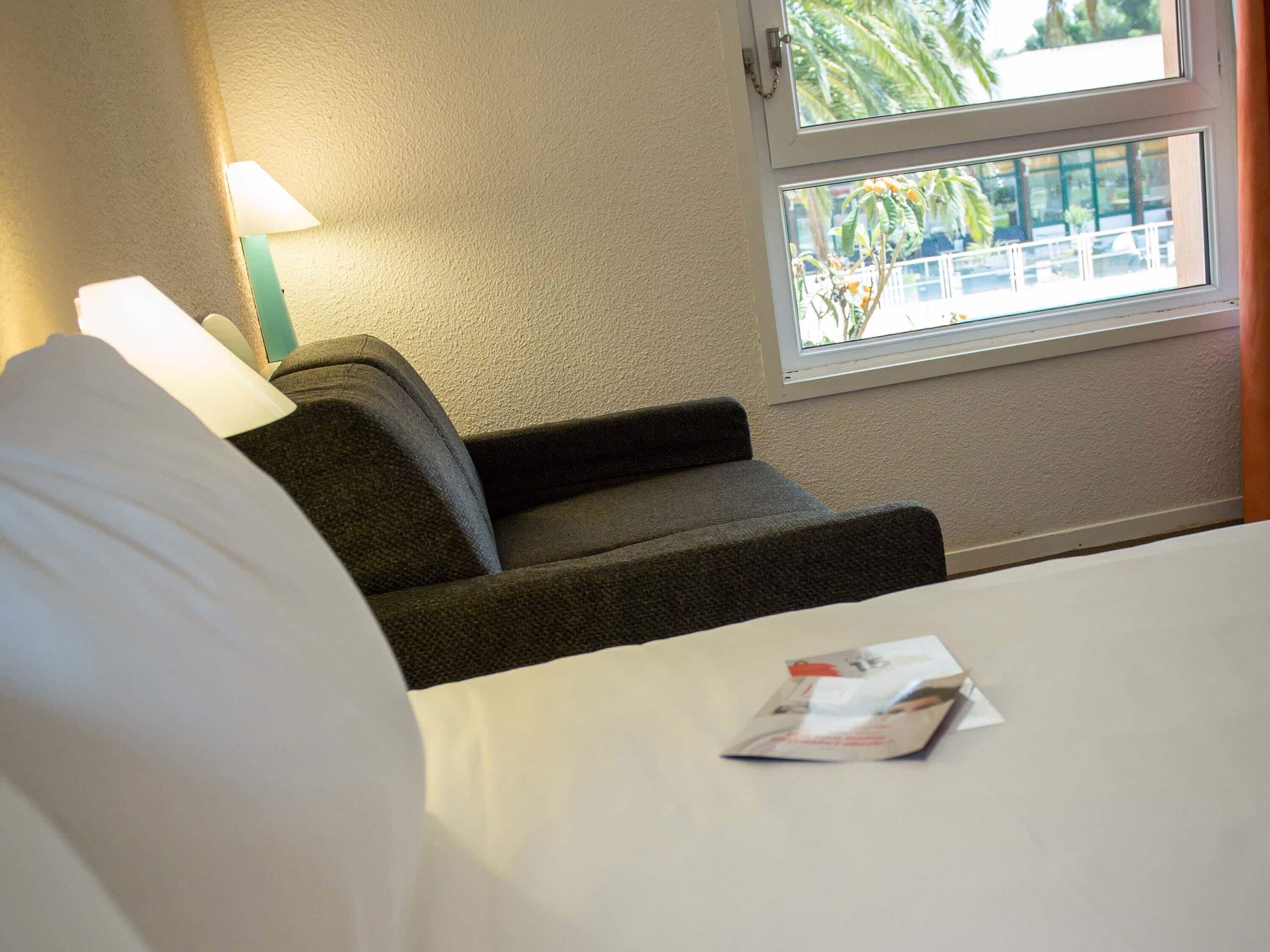Hotellihuone, jossa on sänky, pieni sohva, ikkuna, josta avautuu näkymä palmuille, ja seinässä oleva lamppu.
