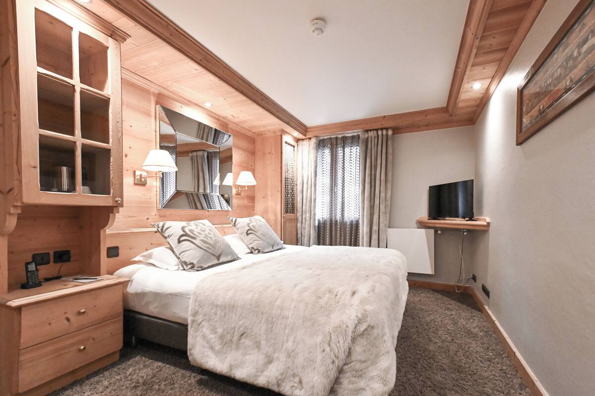 Een slaapkamer met een houten hoofdbord, een groot bed met een zachte witte deken, een nachtkastje met een lamp, een spiegel en een televisie op een plank.