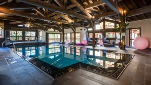 Una piscina interior con techo de madera, paredes de vidrio transparente y una sala iluminada con asientos y una planta.