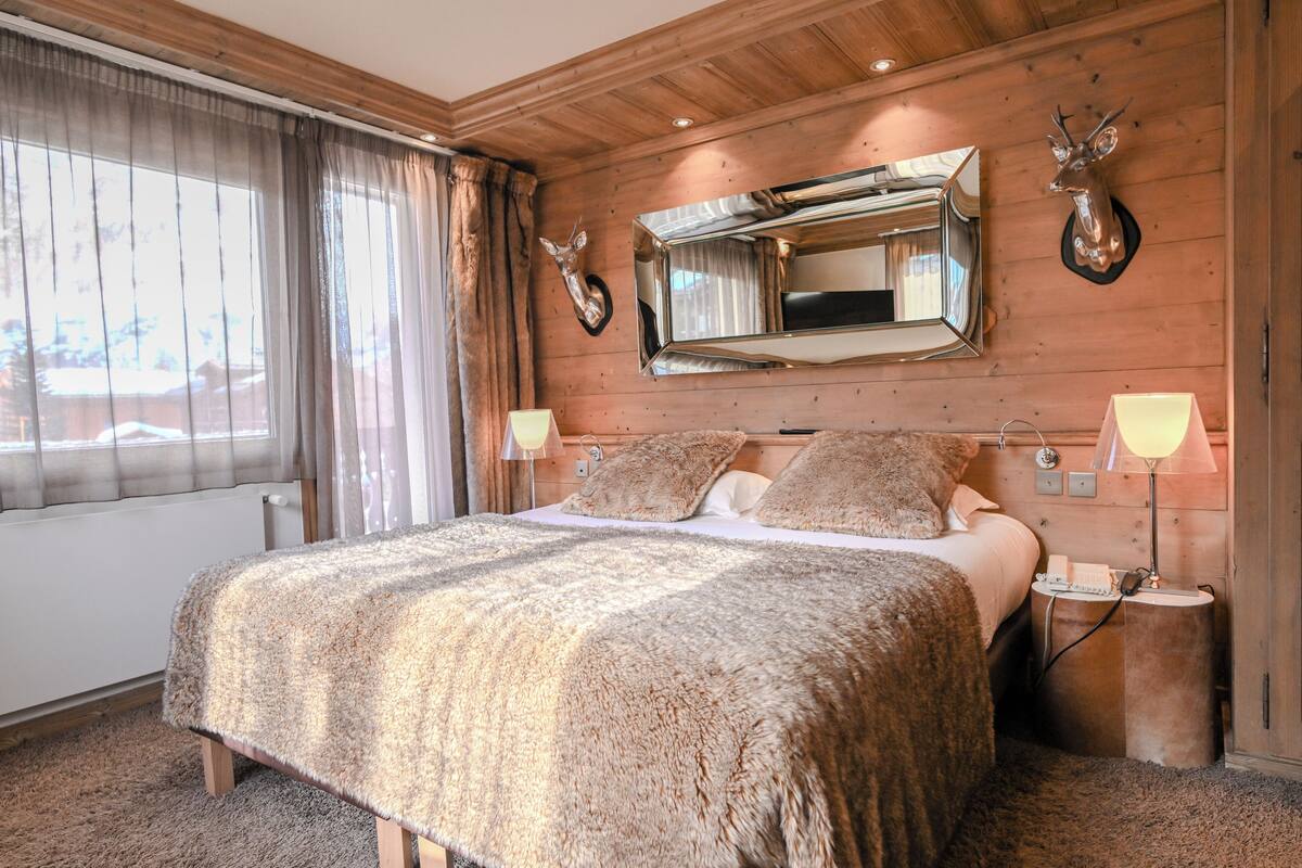 Een slaapkamer met houten lambrisering, een groot bed, twee bedlampen, een vastgeplakte spiegel en een hertenkop aan de muur.